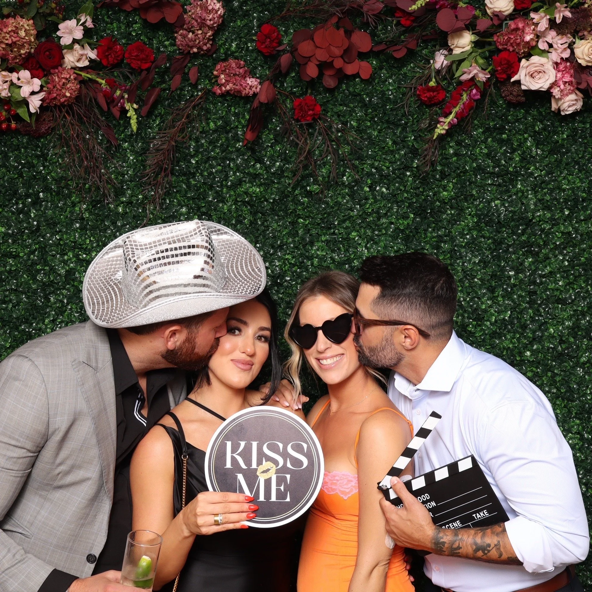 Four people at a photo booth with a green leafy backdrop and pink flowers, one holding a sign that says 'KISS ME', two kissing her cheeks, all smiling, wearing casual and fun accessories.