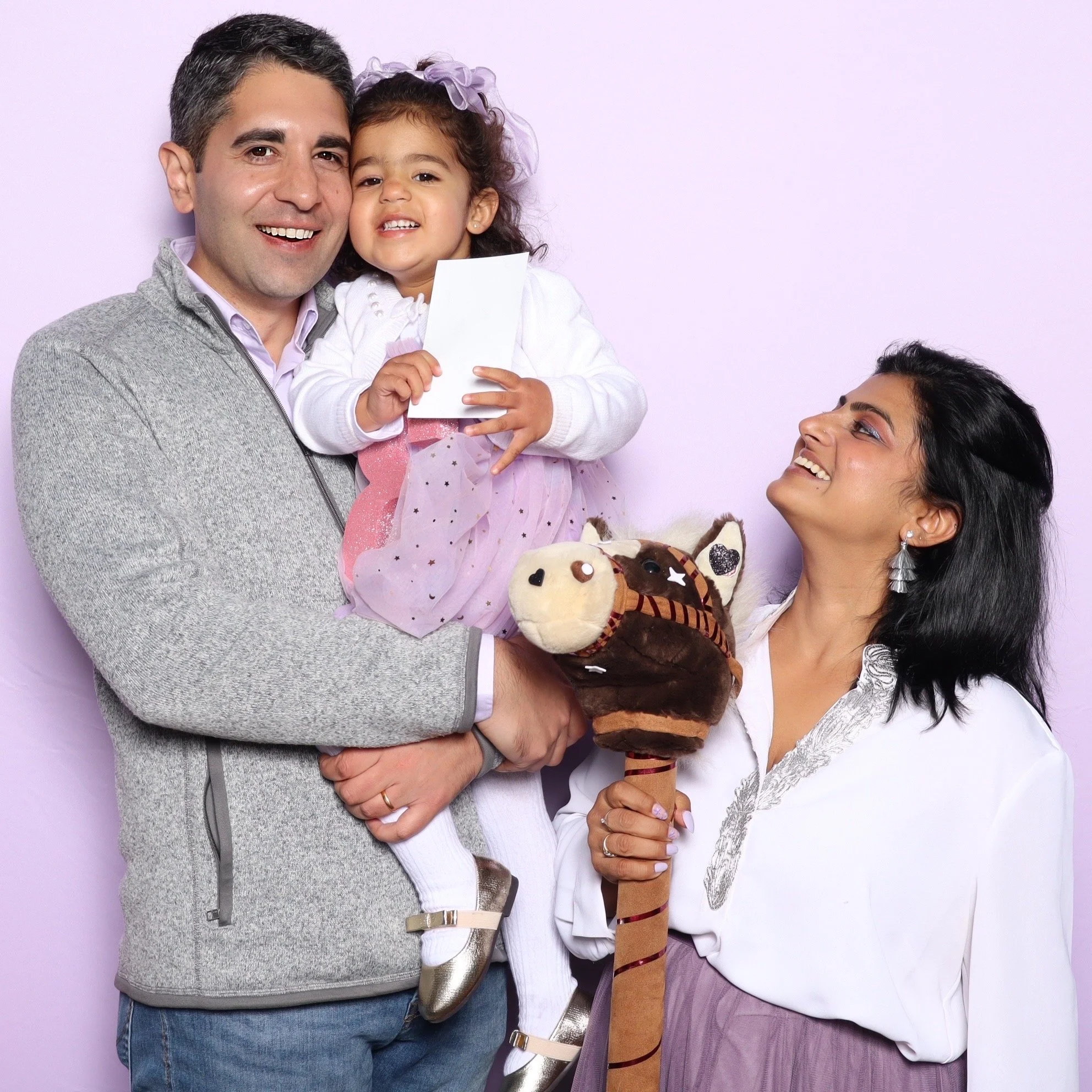 A family celebrating with a young girl, accompanied by her parents, holding a toy giraffe and a gift, against a light purple background.
