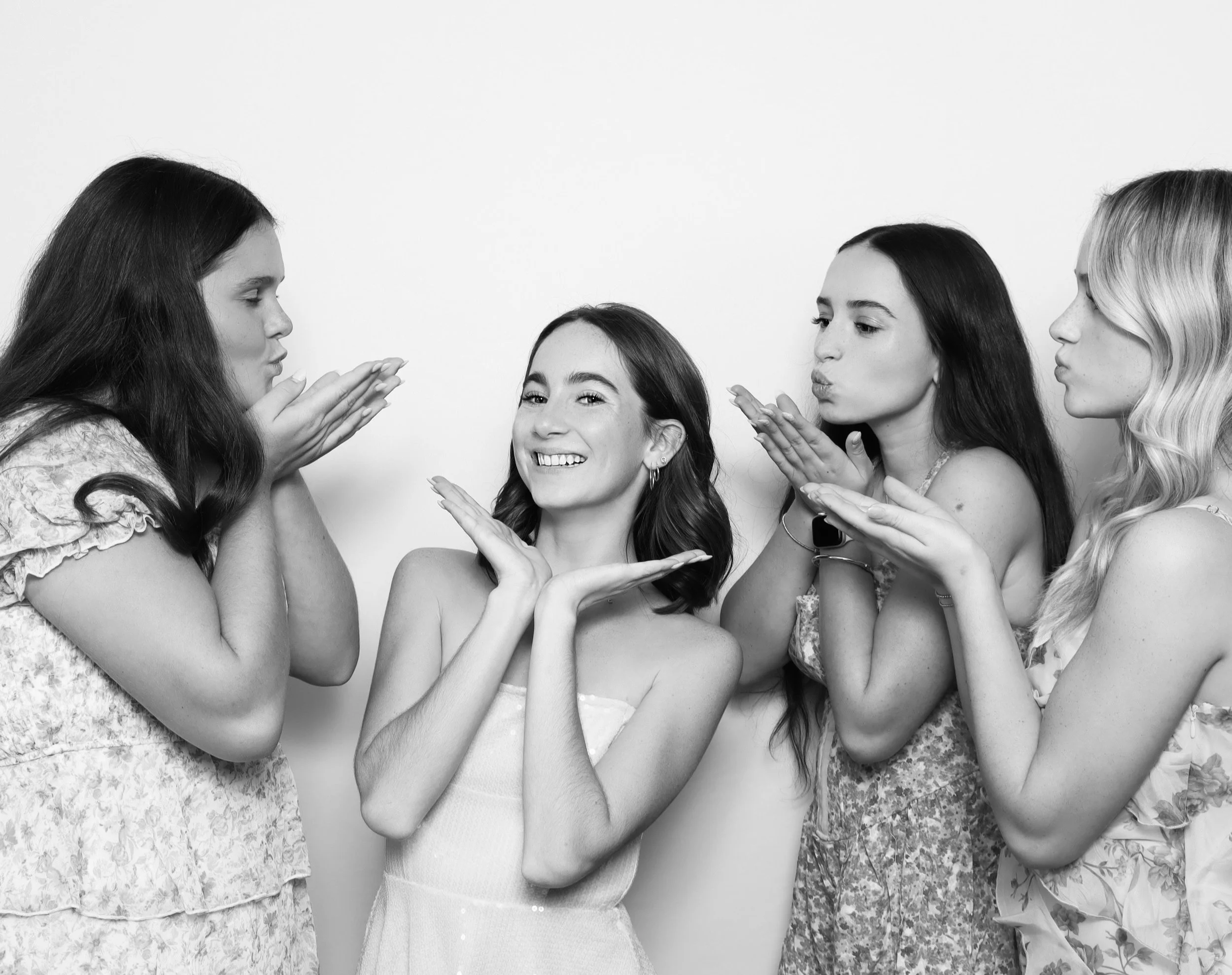 Four women with long hair standing close together, one smiling and surrounded by the other three who are blowing kisses towards her, in a black and white photo.