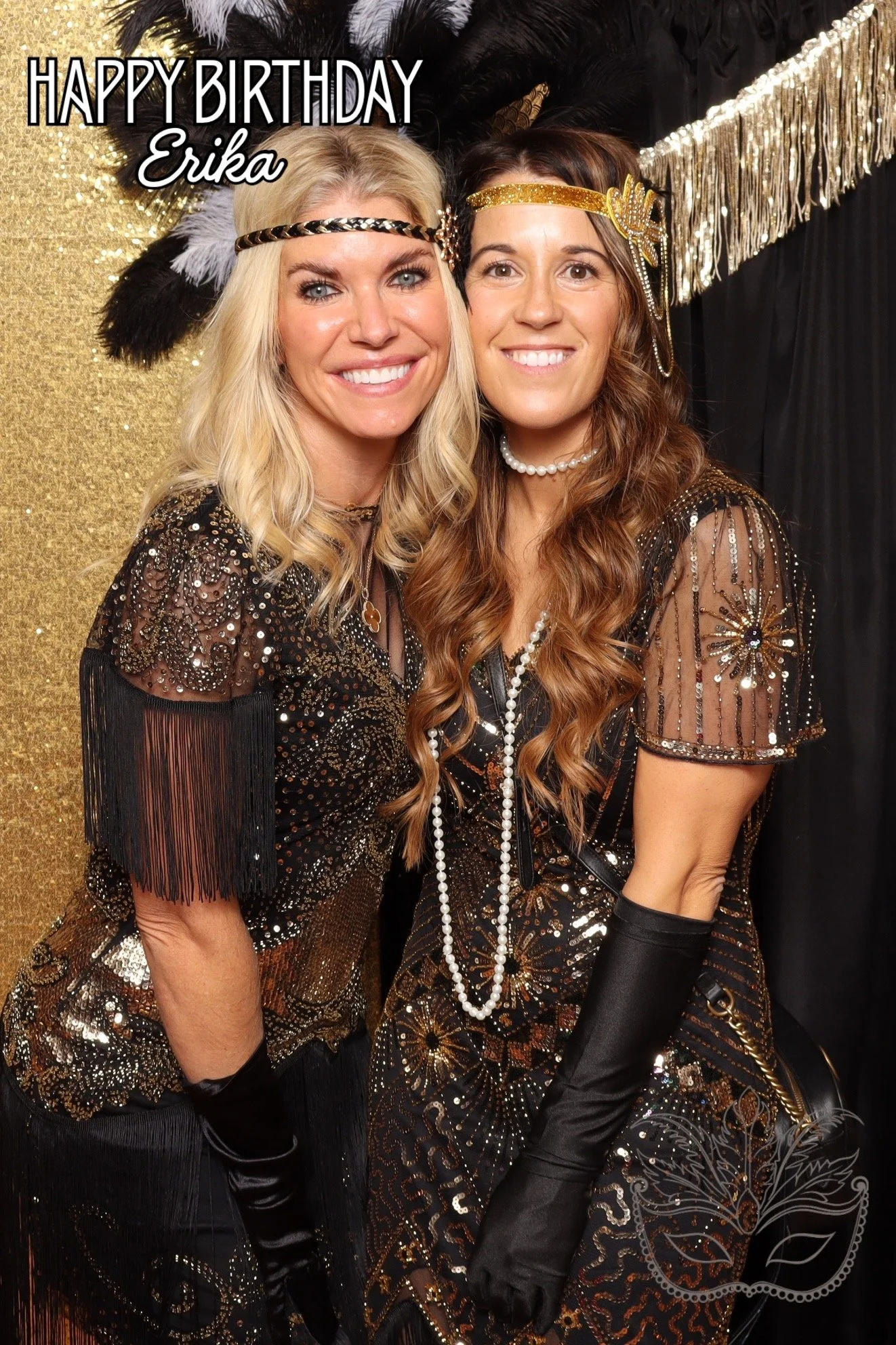Two women dressed in black and gold costumes with sequins, beads, and fringe, smiling for a photo at a birthday celebration. The background has black and gold decorations, including feathers and fringe, with a sign that reads "Happy Birthday Erika." 