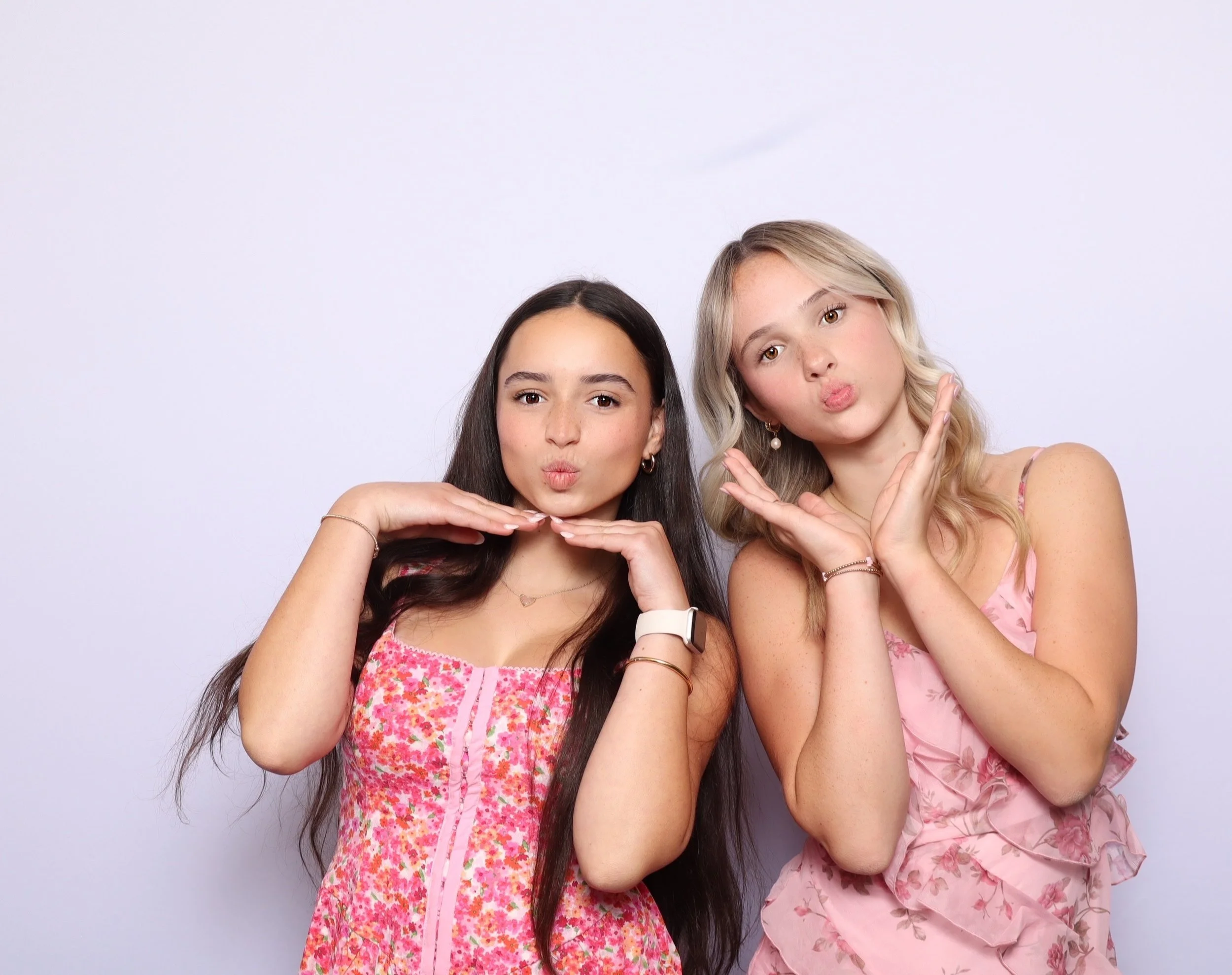 Two young women in pink dresses posing with playful kissy faces and hand gestures in front of a plain light background.