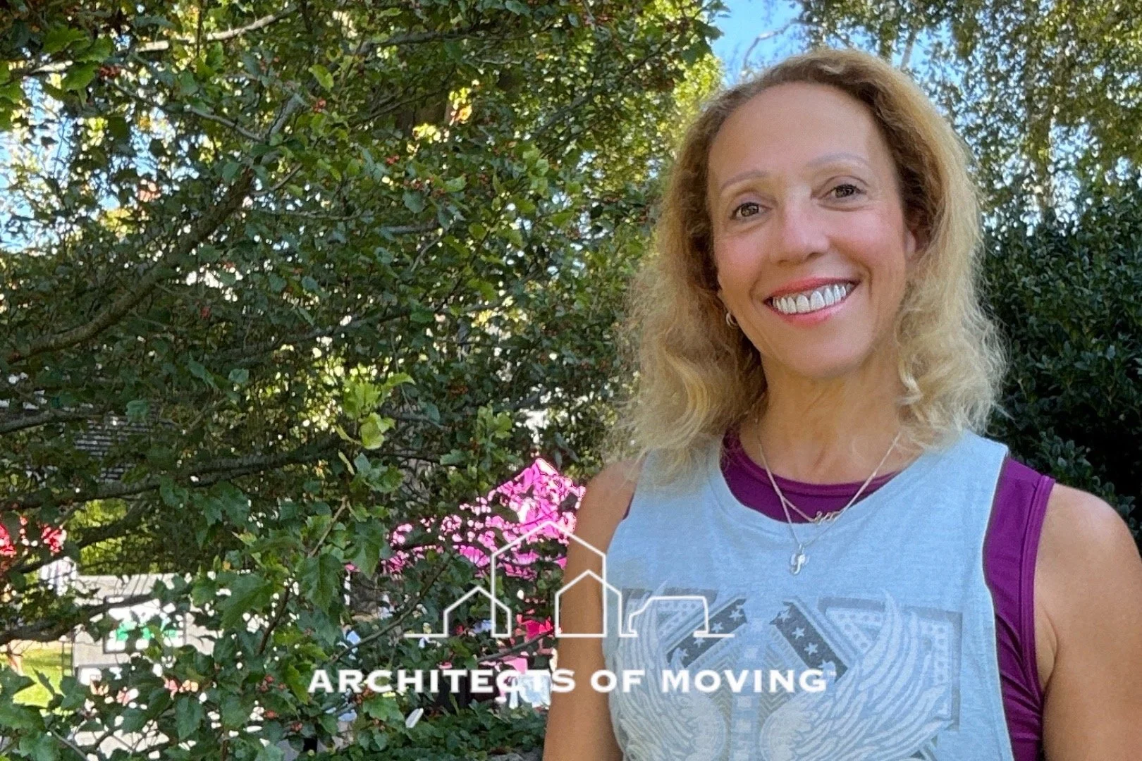 A woman with curly blonde hair smiling outdoors in front of trees and greenery, wearing a tank top and jewelry, with a logo for architects of moving.
