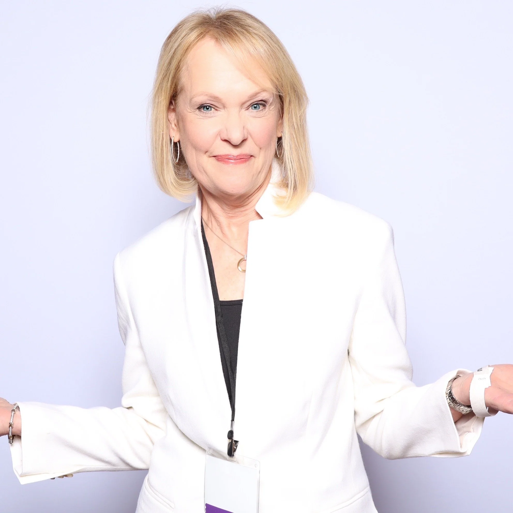 A woman with shoulder-length blonde hair, wearing a white blazer over a black top, smiles with her arms outstretched against a plain light background.