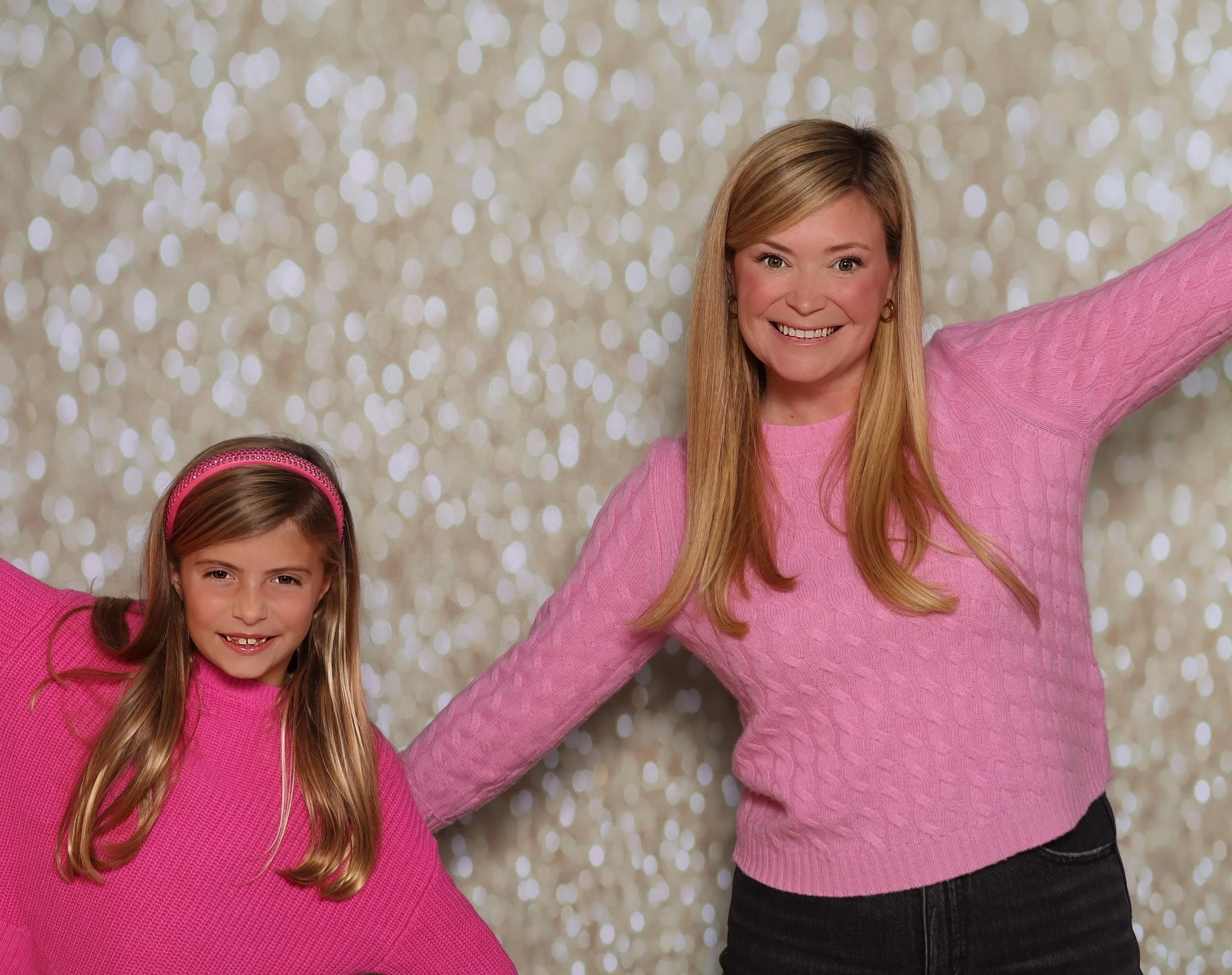 A woman and a young girl in pink sweaters smiling and posing with outstretched arms against a sparkly, bokeh background.
