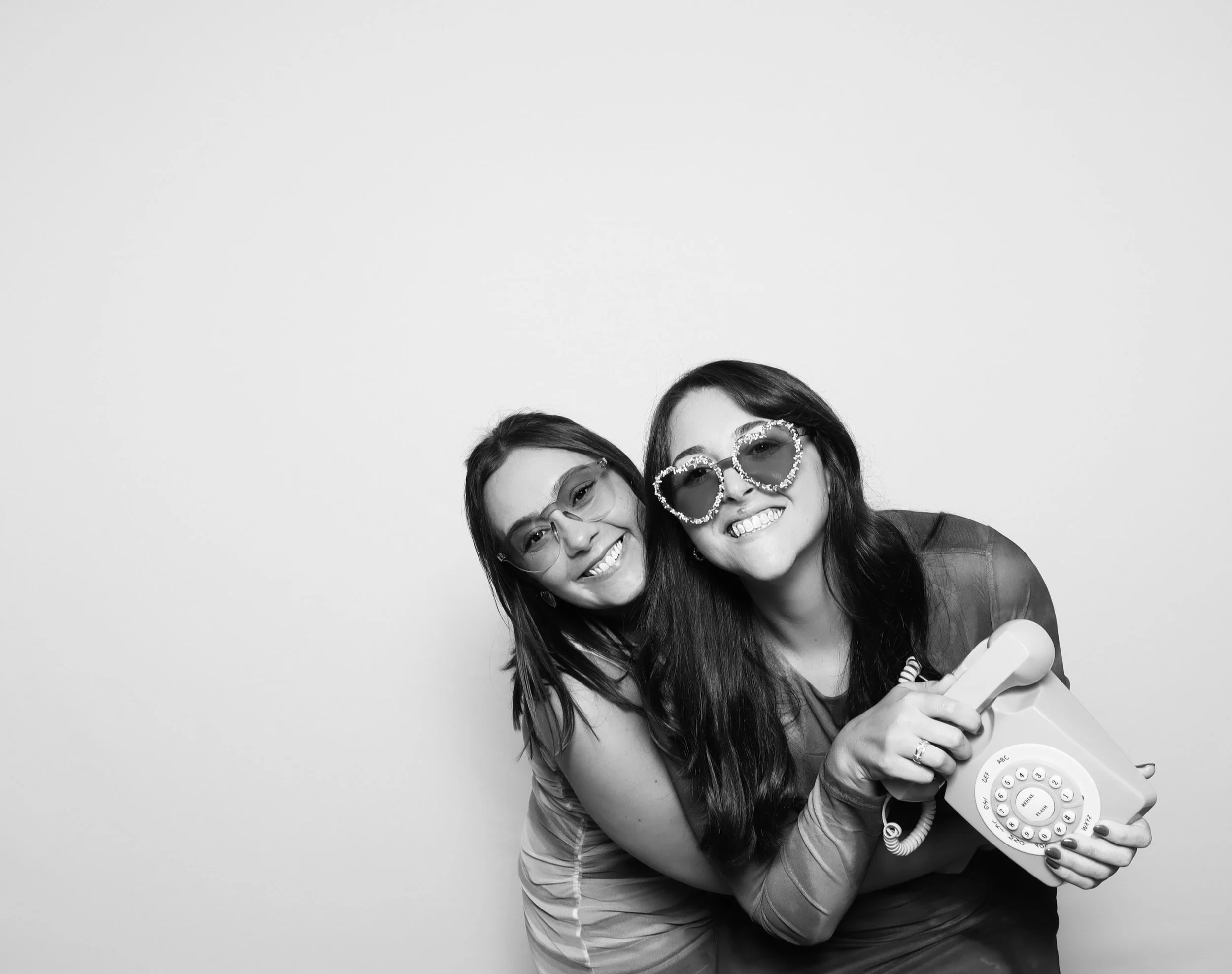 Two women smiling, wearing sunglasses, one with heart-shaped glasses, holding a vintage rotary phone, against a plain light-colored background.