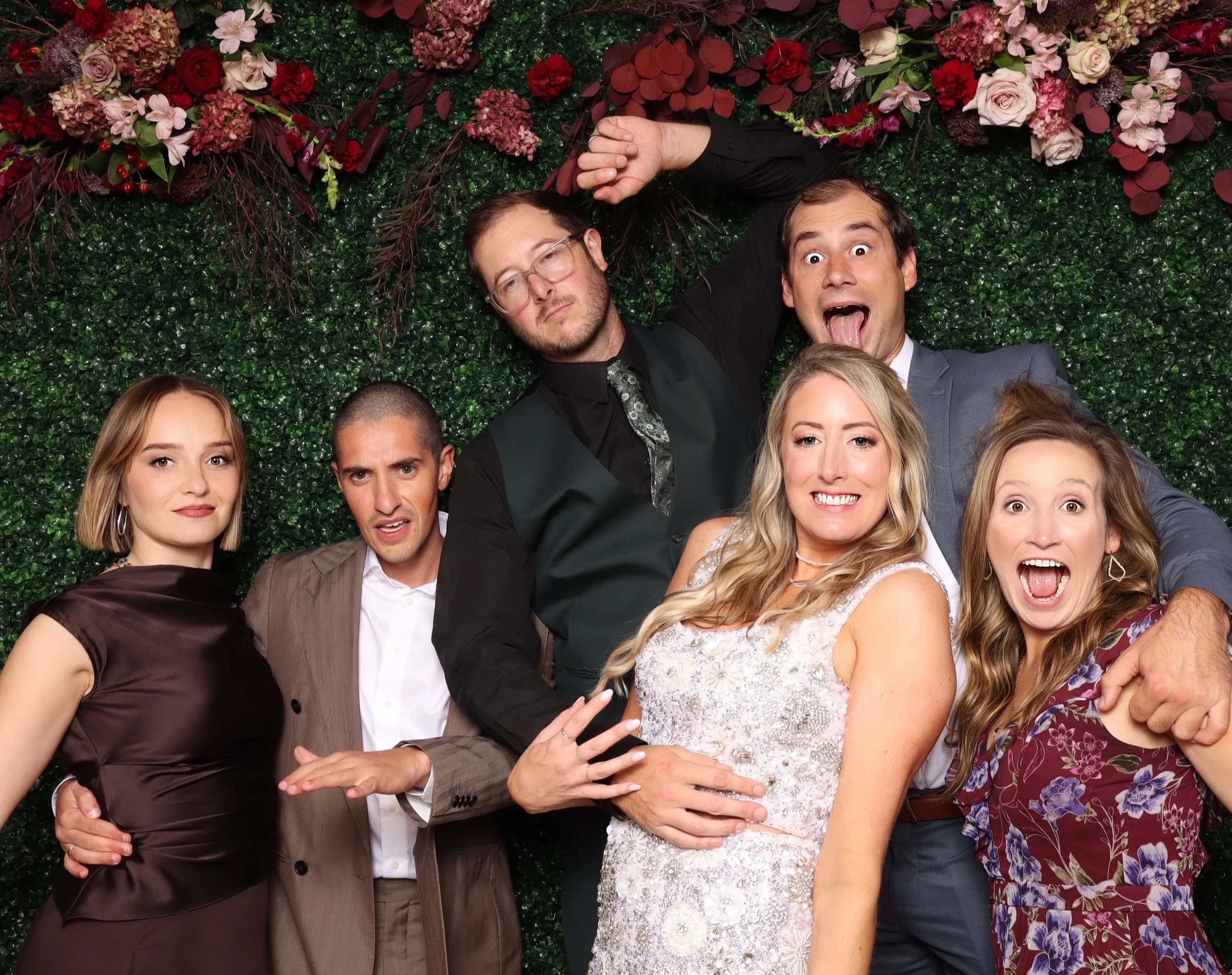 Group of six people posing and making funny faces together in front of a green leafy backdrop decorated with pink, red, and purple flowers.