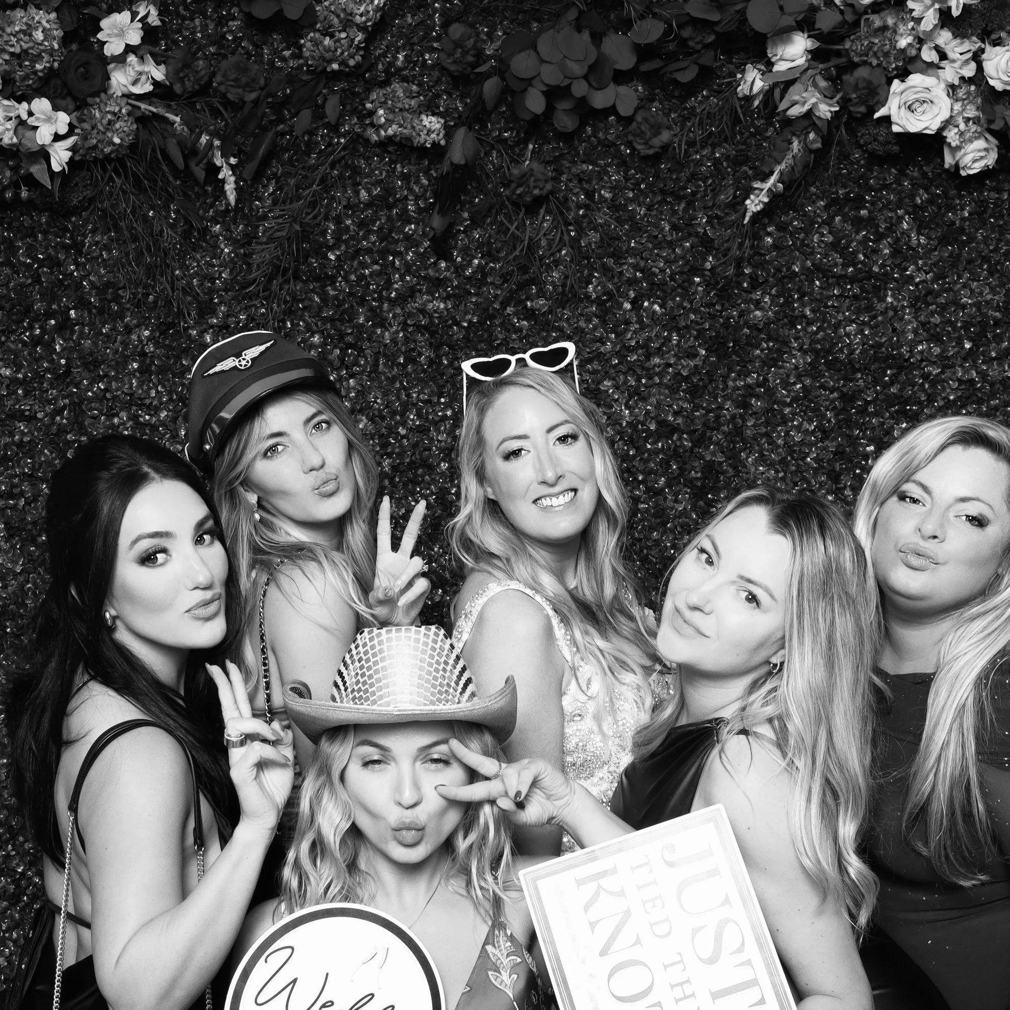 Six women posing for a photo in front of a floral backdrop, with some making peace signs or puckering lips, some wearing fun accessories like glasses, hats, and a police cap.