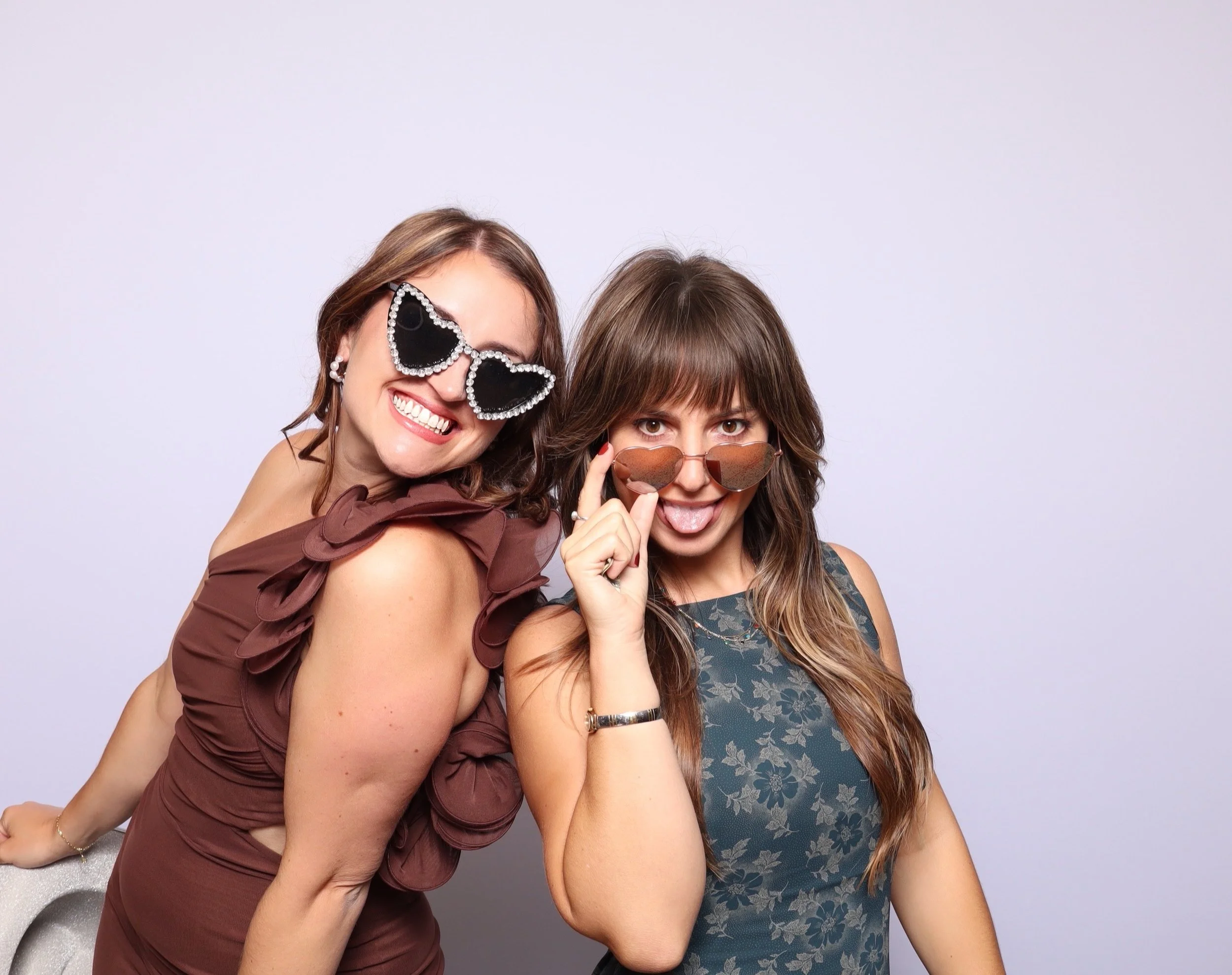 Two women posing playfully, wearing sunglasses and trendy dresses, against a plain background.