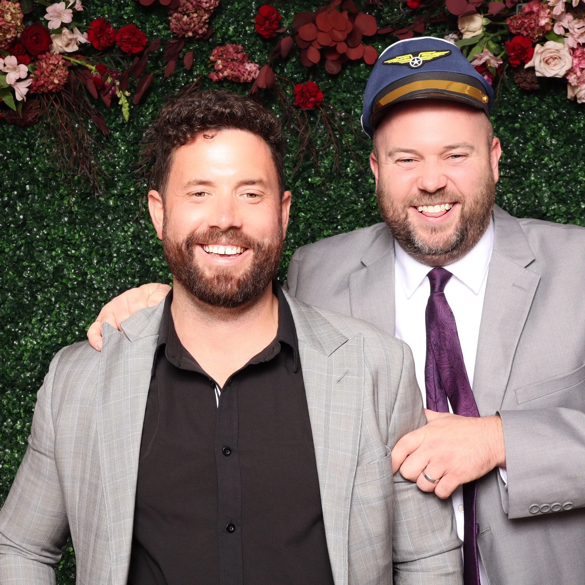 Two men smiling, one with a beard wearing a black shirt and grey blazer, the other also with a beard, wearing a grey suit, purple tie, and a pilot's hat, in front of a floral backdrop.