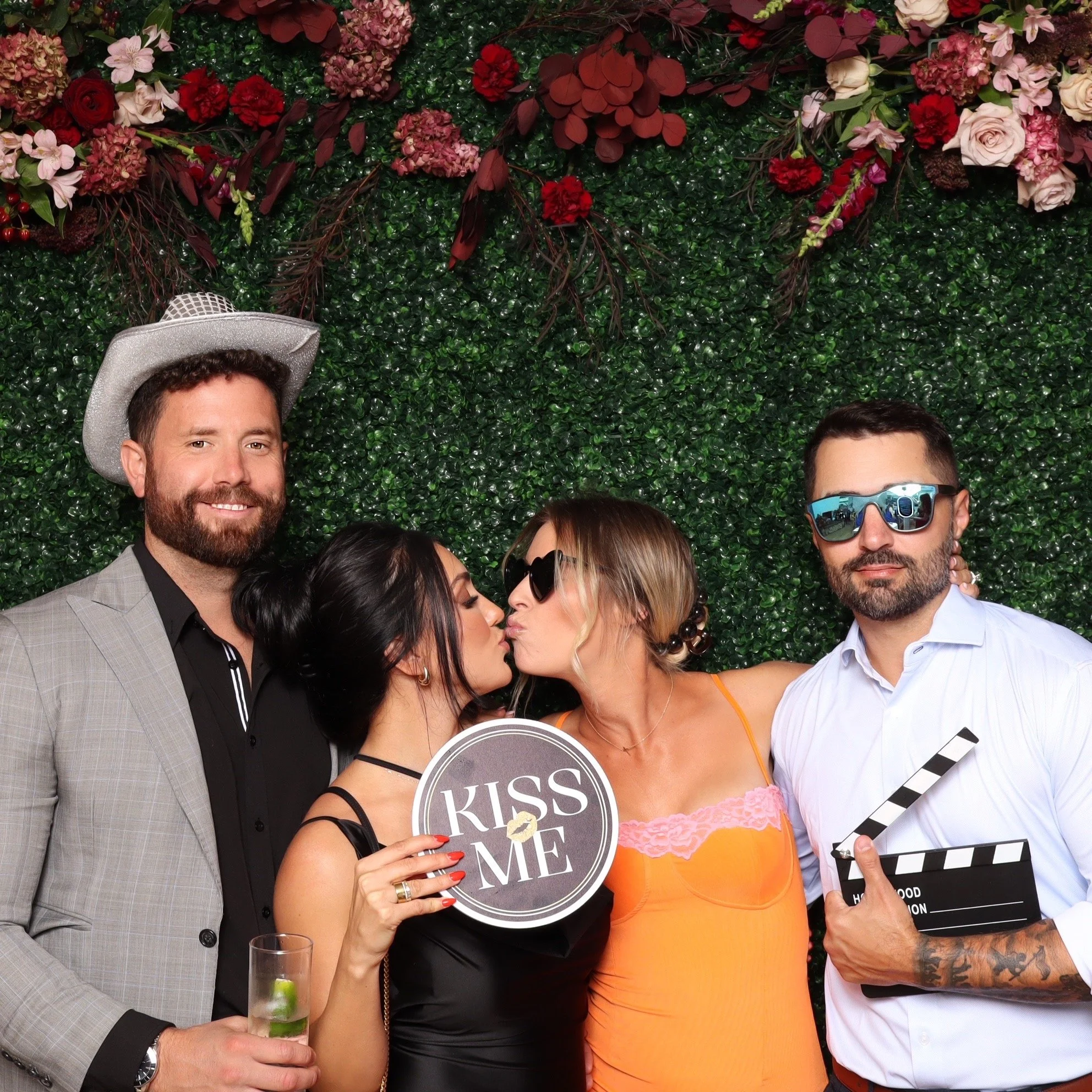Four people at a photo booth, with a green leafy backdrop and pink, red, and white flowers. One man in a gray suit and cowboy hat, holding a drink, smiles at the camera. Two women and another man are in the middle; the women are kissing, with one wom