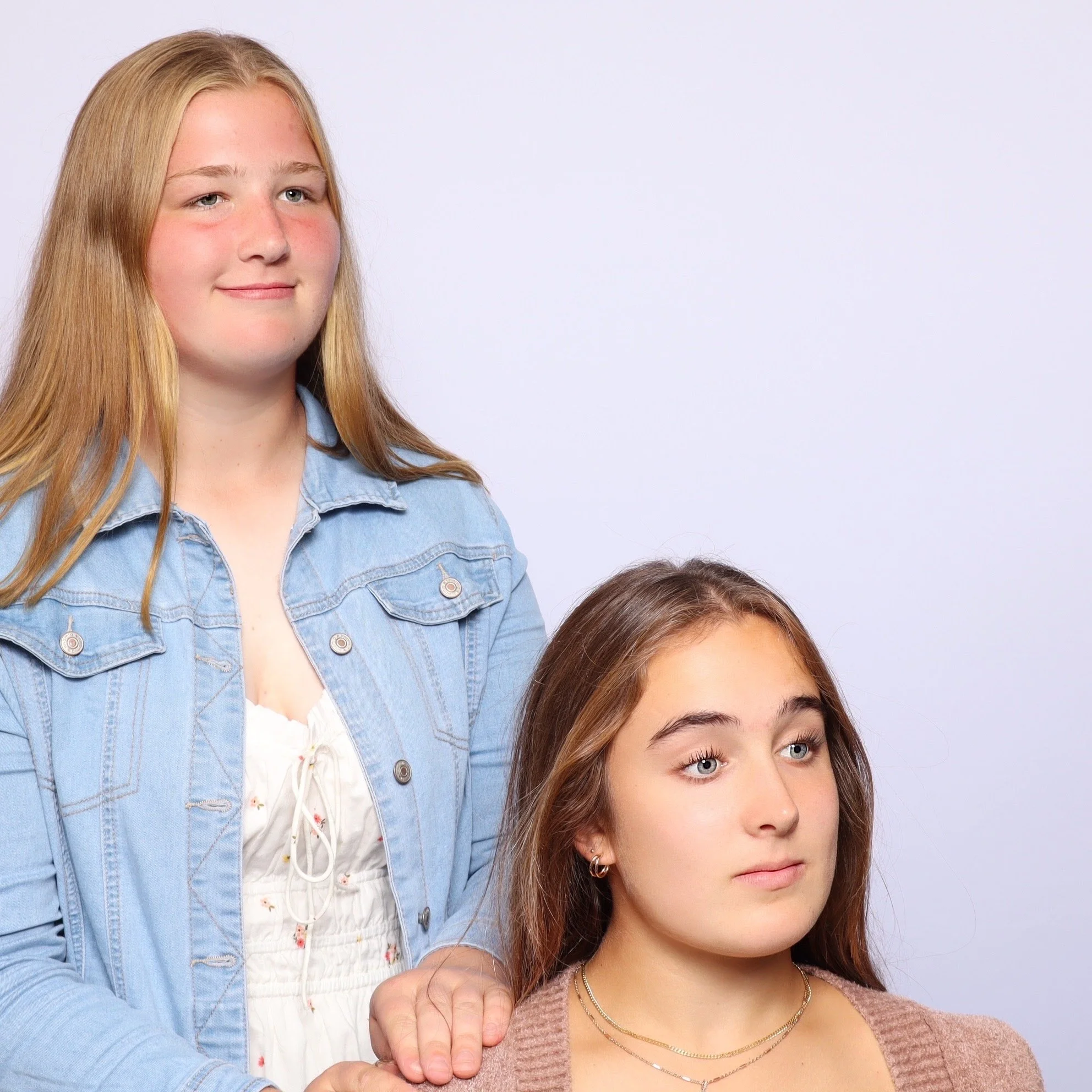 Two young women, one with blonde hair in a denim jacket and white top, and one with brown hair in a brown sweater and jewelry, pose against a plain light background.