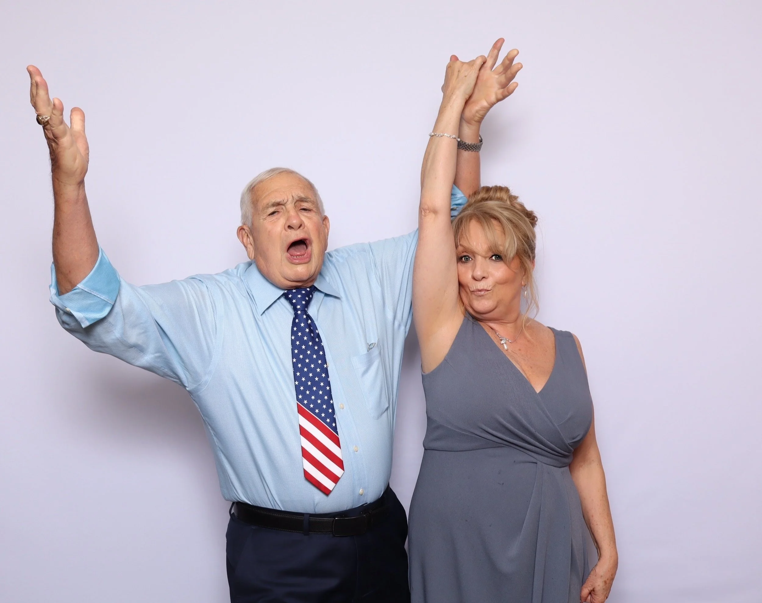 A joyful elderly couple with the man raising his hands in excitement and the woman smiling, standing against a plain white background.