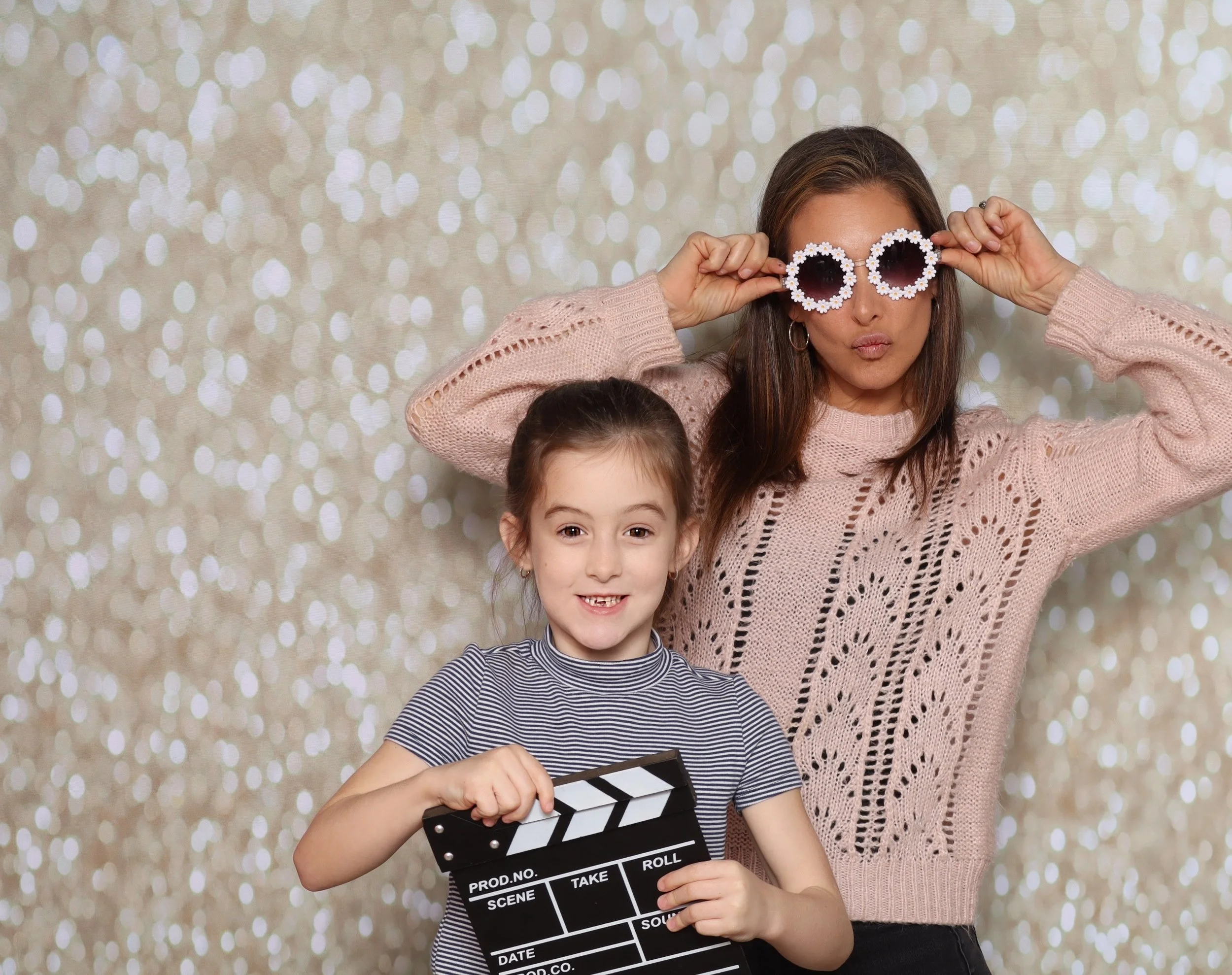 A young girl holding a film slate with a big smile, and an adult woman behind her making funny sunglasses with daisy-shaped frames, both standing in front of a sparkly, bokeh background.