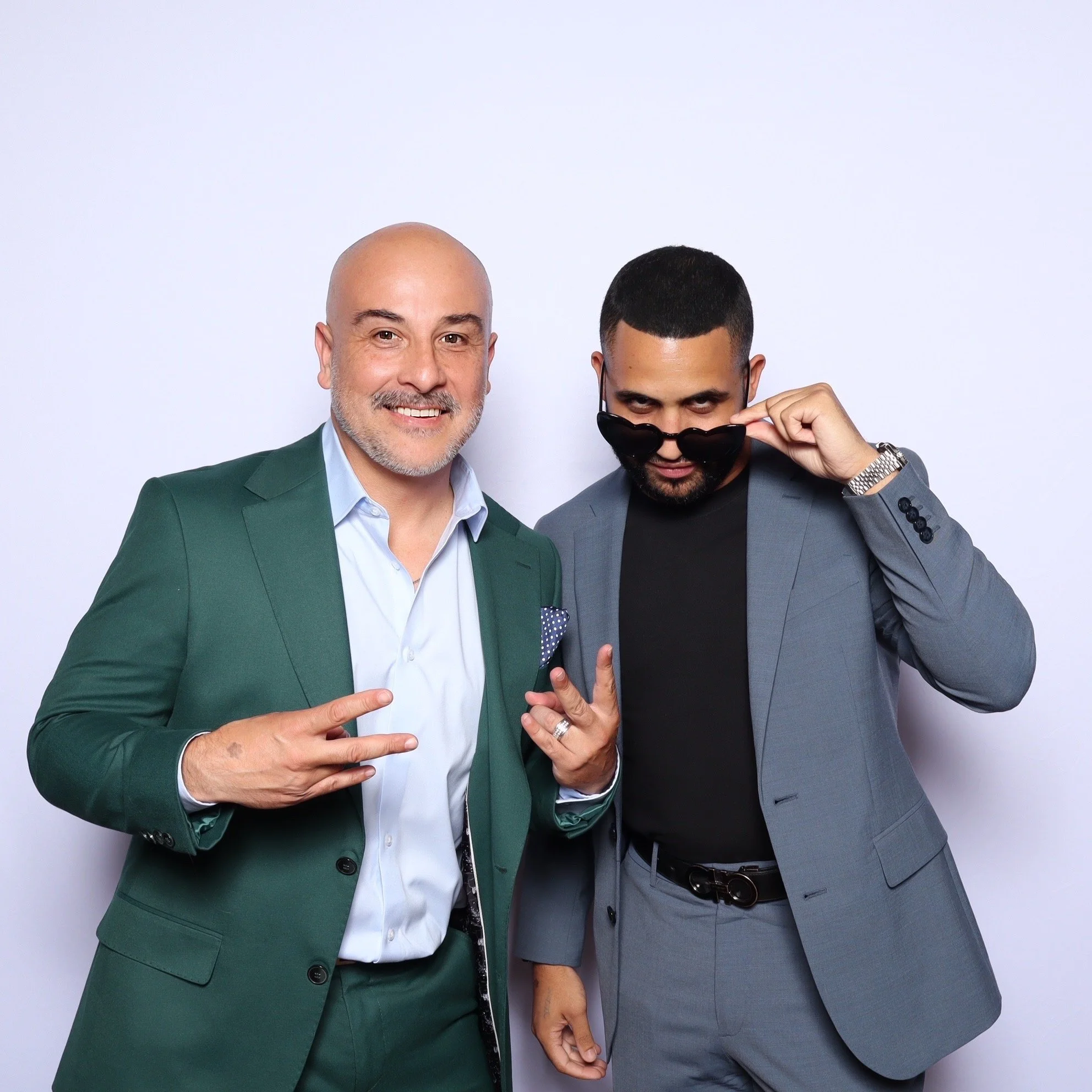 Two men in suits posing for the camera with confident expressions against a plain white background. One man points with his fingers while the other adjusts sunglasses.