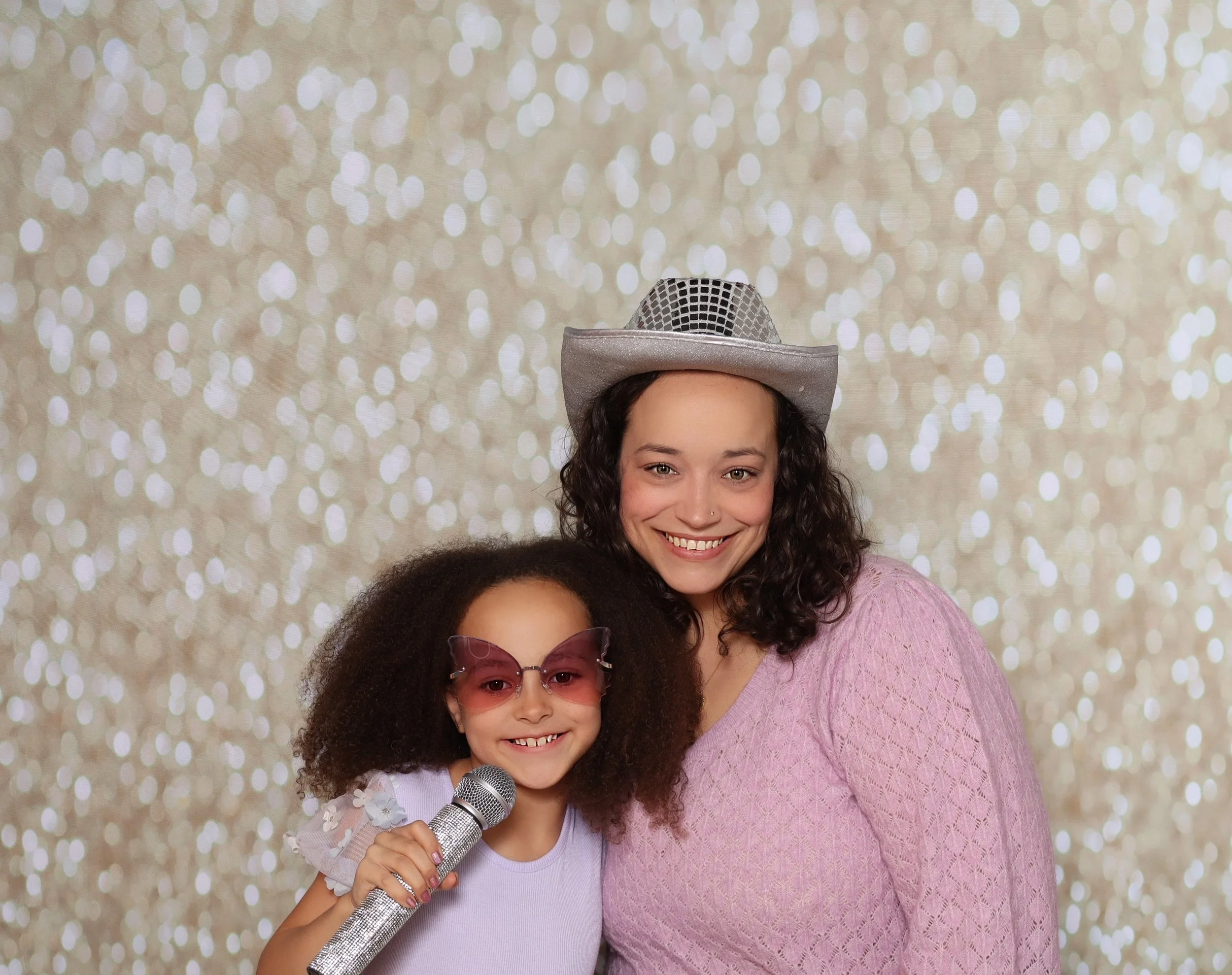 Two women smiling, one wearing pink sweater and silver cowboy hat, the other with sunglasses and microphone, against a sparkly background.