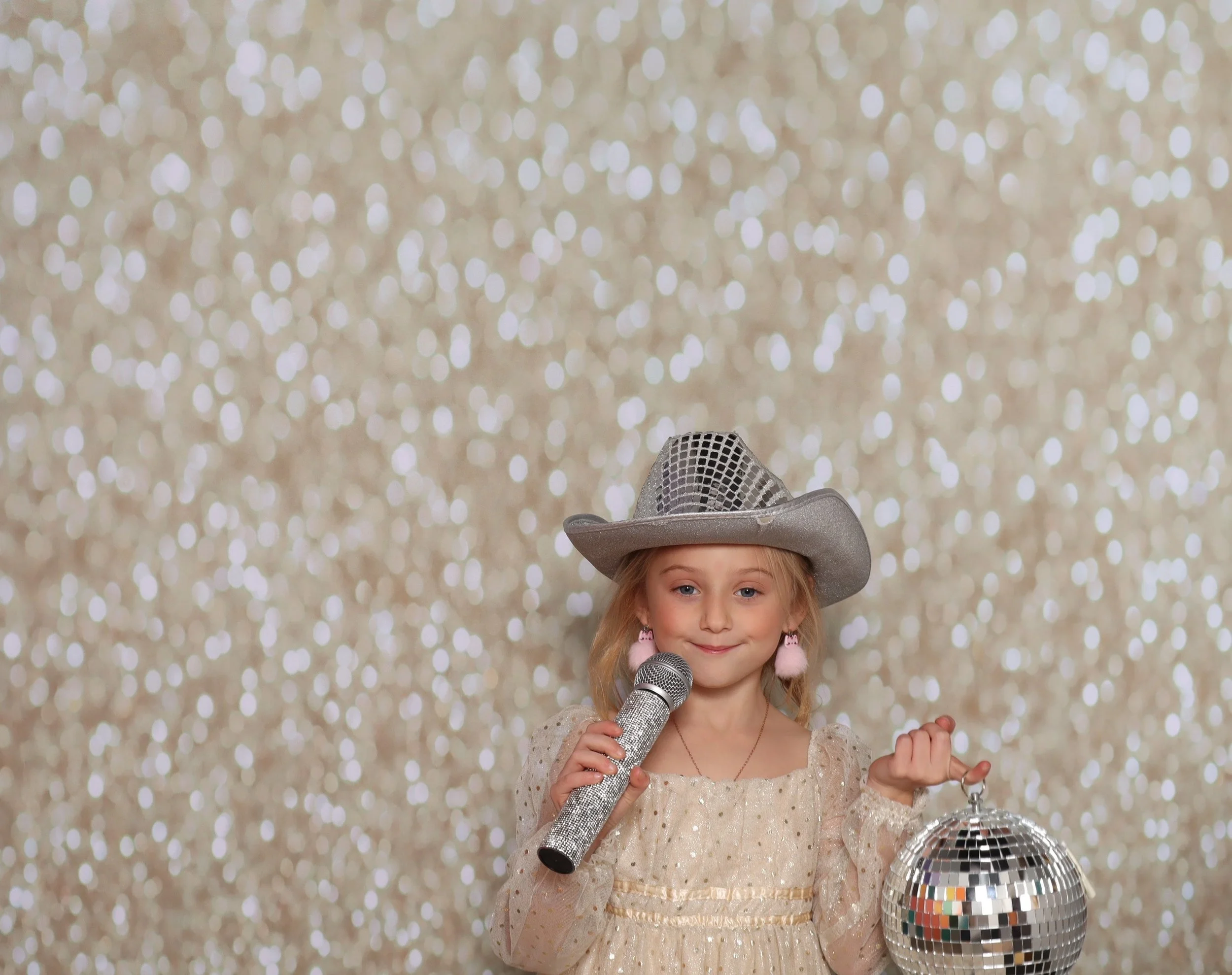 A young girl wearing a sparkly dress and a cowboy hat, holding a microphone and a small disco ball, standing in front of a glittery gold background.