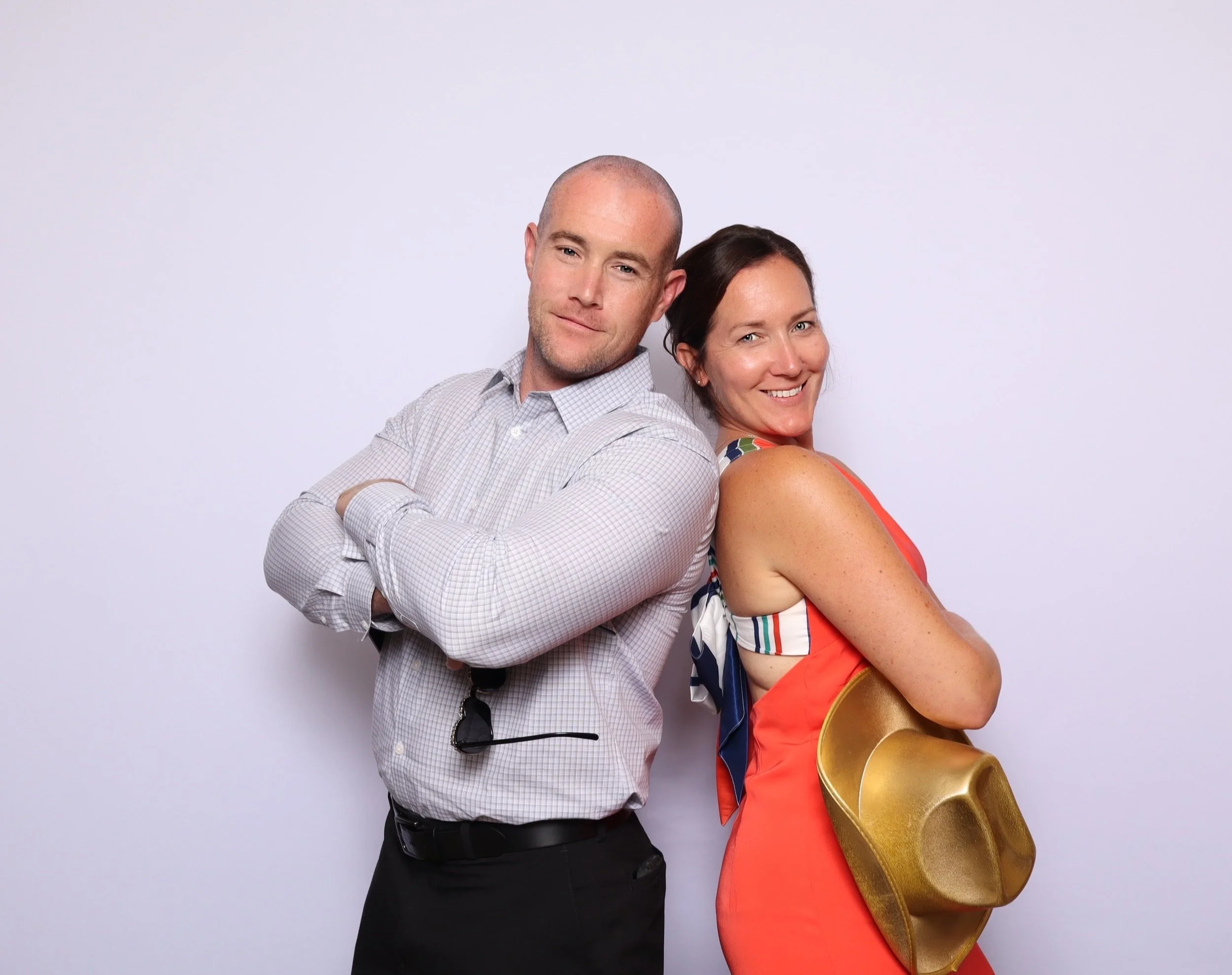 A man and woman stand back-to-back, smiling with arms crossed against a plain white background. The man wears a light checked shirt and black pants. The woman wears a bright orange sleeveless dress and holds a shiny gold cowboy hat, with a multicolor