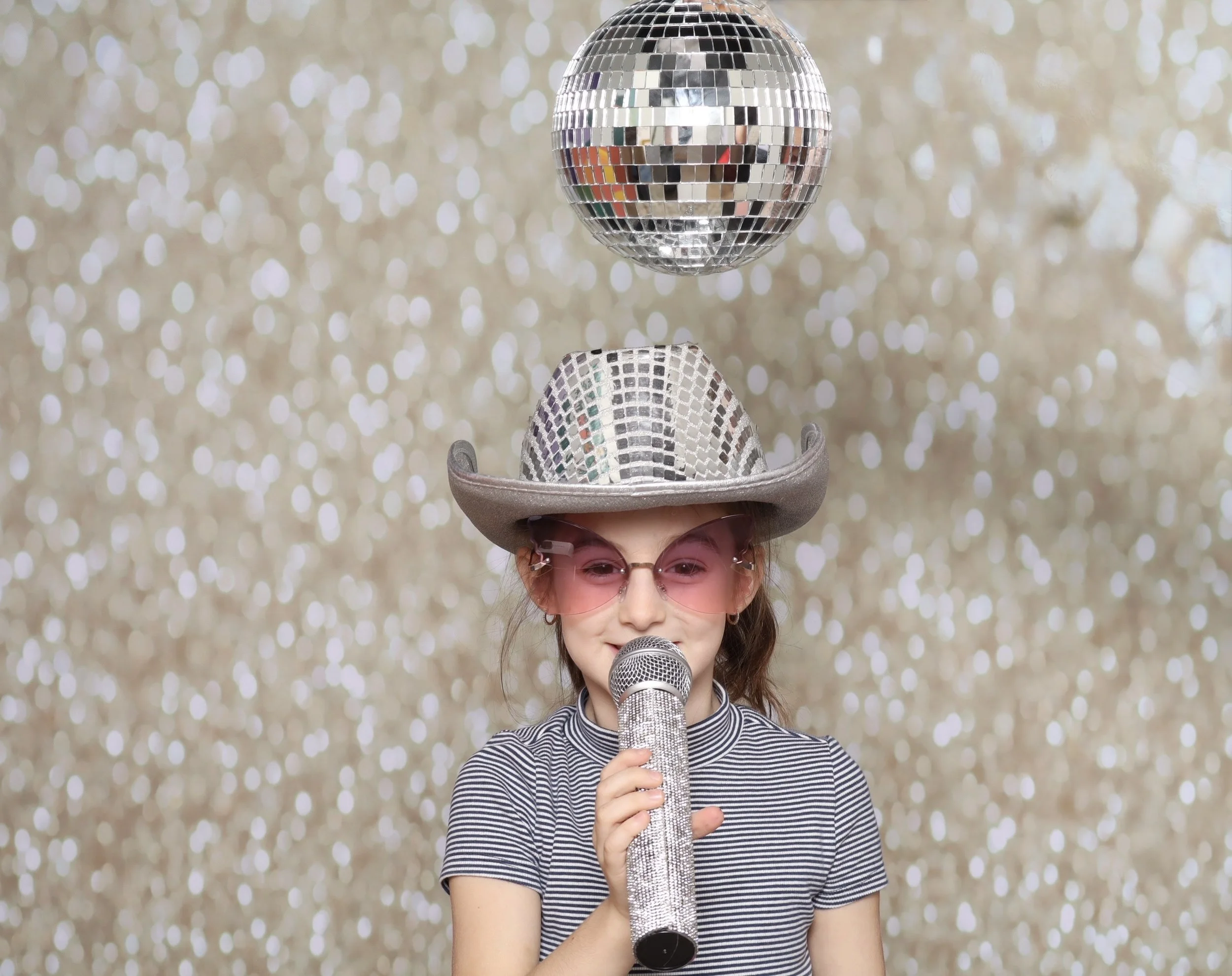 Young girl dressed in a striped shirt, wearing a cowboy hat and pink sunglasses, holding a glittery microphone, with a disco ball above her head against a glittery background.