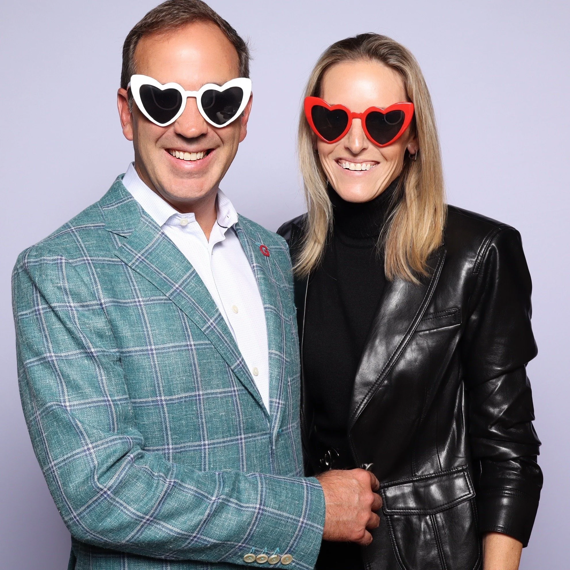 A man and woman smiling, wearing heart-shaped sunglasses, with the man in a plaid suit and the woman in a black leather jacket.