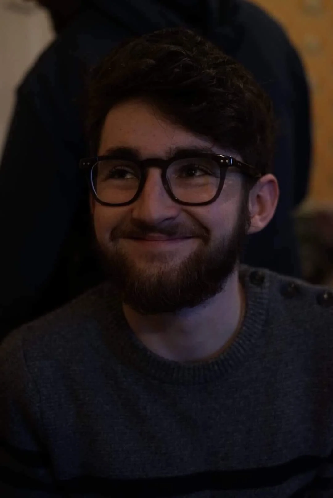 A smiling young man with dark hair, beard, and glasses, resting on a dark surface.