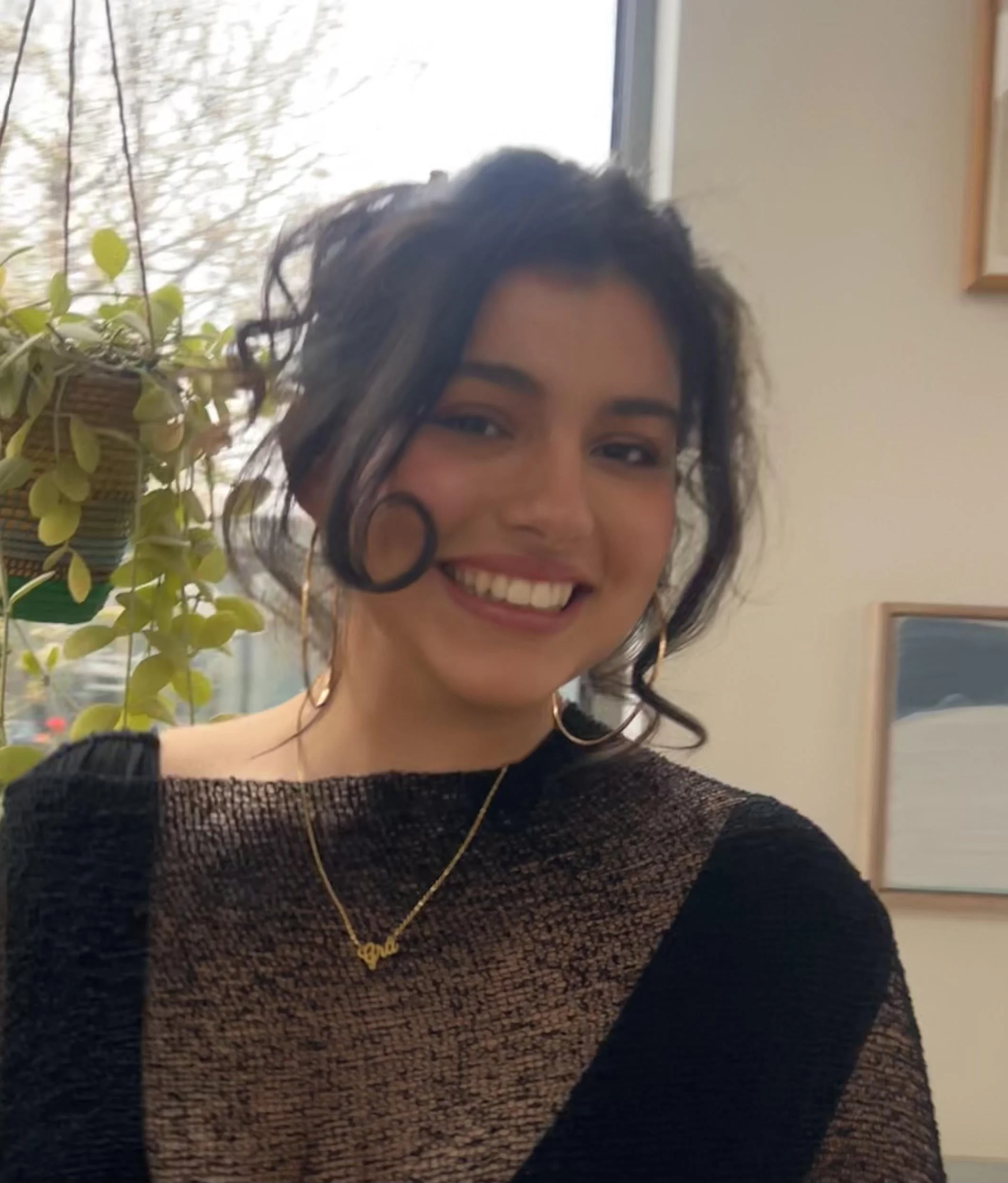 A smiling woman with curly black hair, wearing a black top, a gold necklace with a name pendant, and hoop earrings, taking a selfie near a window with a potted plant visible on the side.