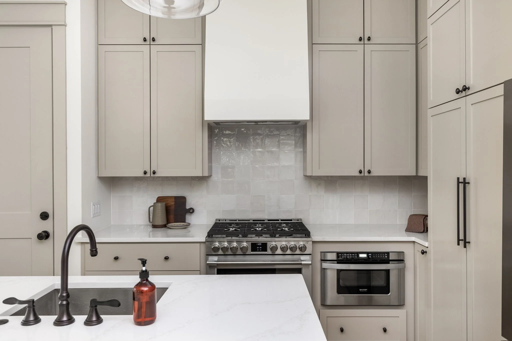 A modern kitchen with white cabinetry, a gas stove with oven, a white tile backsplash, and a white countertop with a dark wood cutting board, plates, and a red soap dispenser near a black faucet.