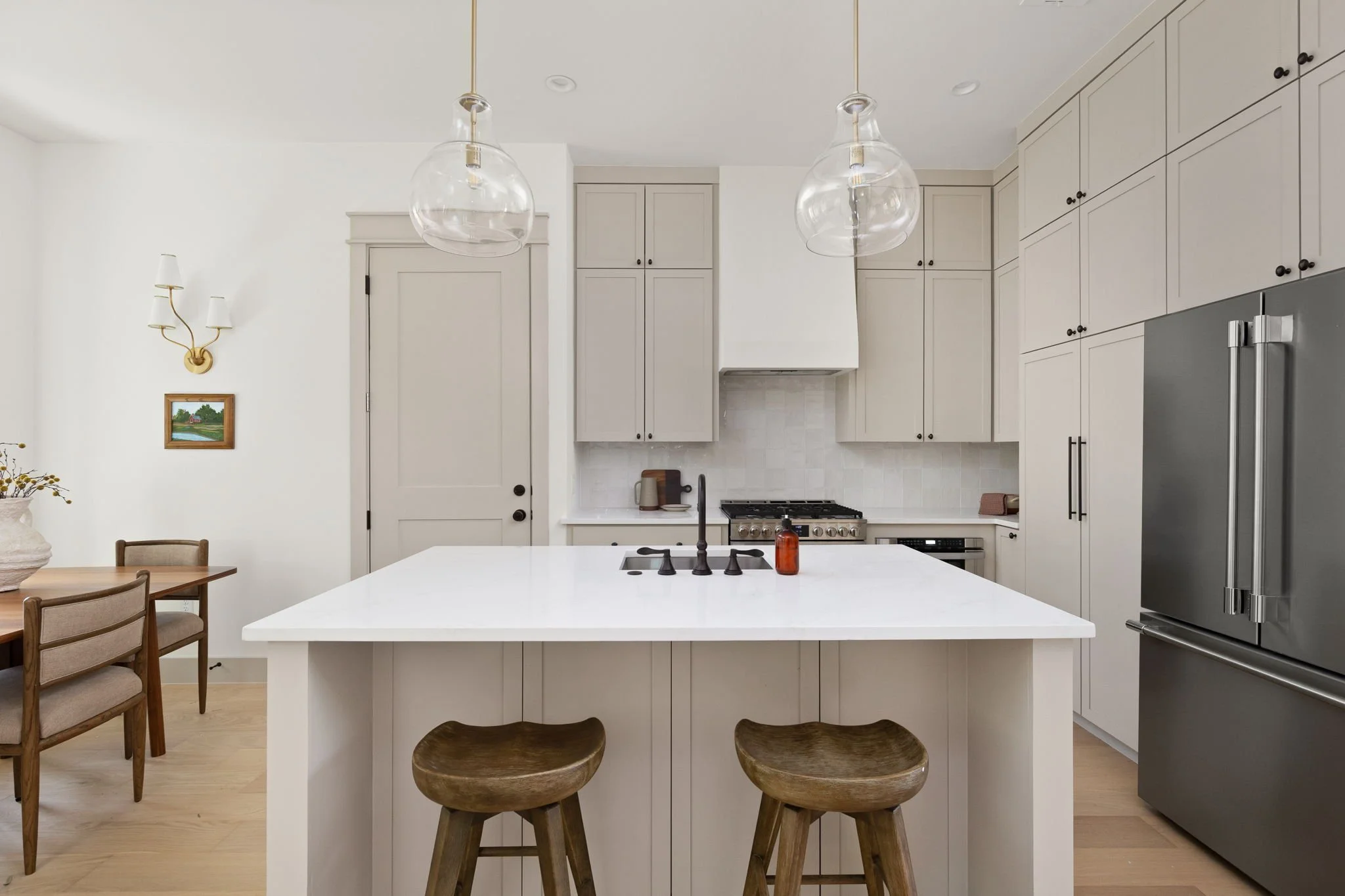 Modern kitchen with white cabinetry, central island with wooden bar stools, stainless steel refrigerator, hanging glass pendant lights, and a white tiled backsplash.