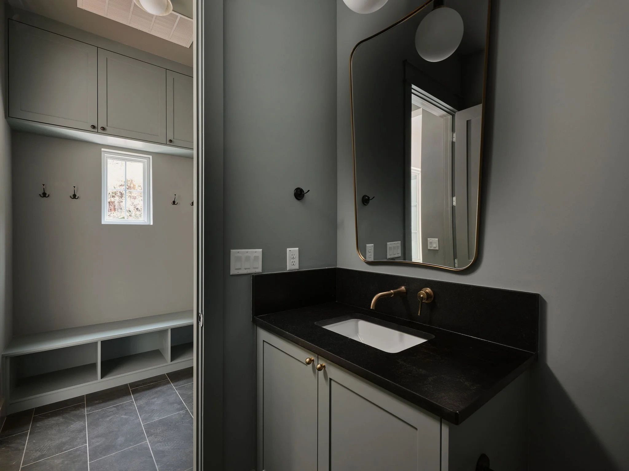 A modern bathroom with a black countertop sink, a large mirror with a gold frame, gray walls, and a closet area with hooks and storage space.
