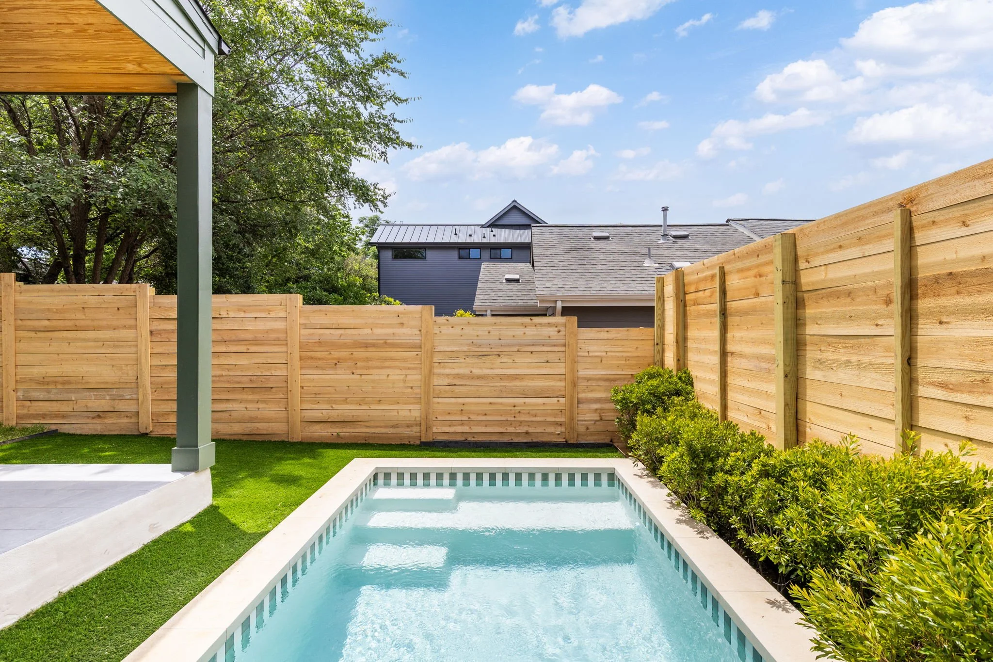 Backyard with a swimming pool, green grass, bushes, a wooden privacy fence, and houses with gray roofs in the background under a blue sky with clouds.