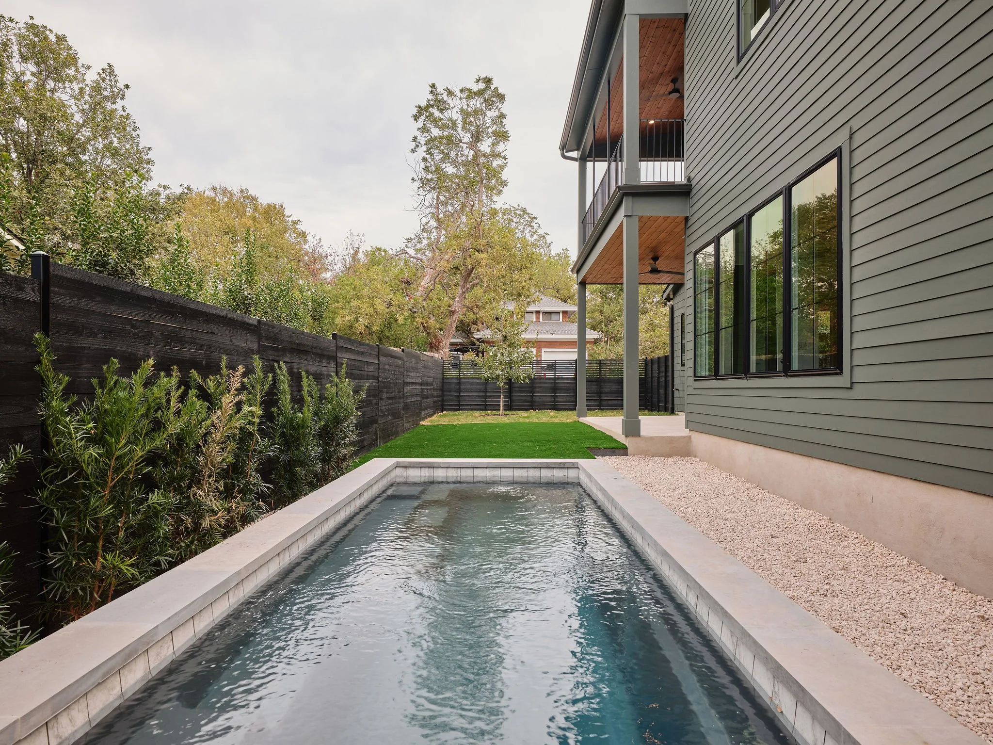 Backyard with small rectangular pool, green lawn, black privacy fence, and modern gray house with large windows.