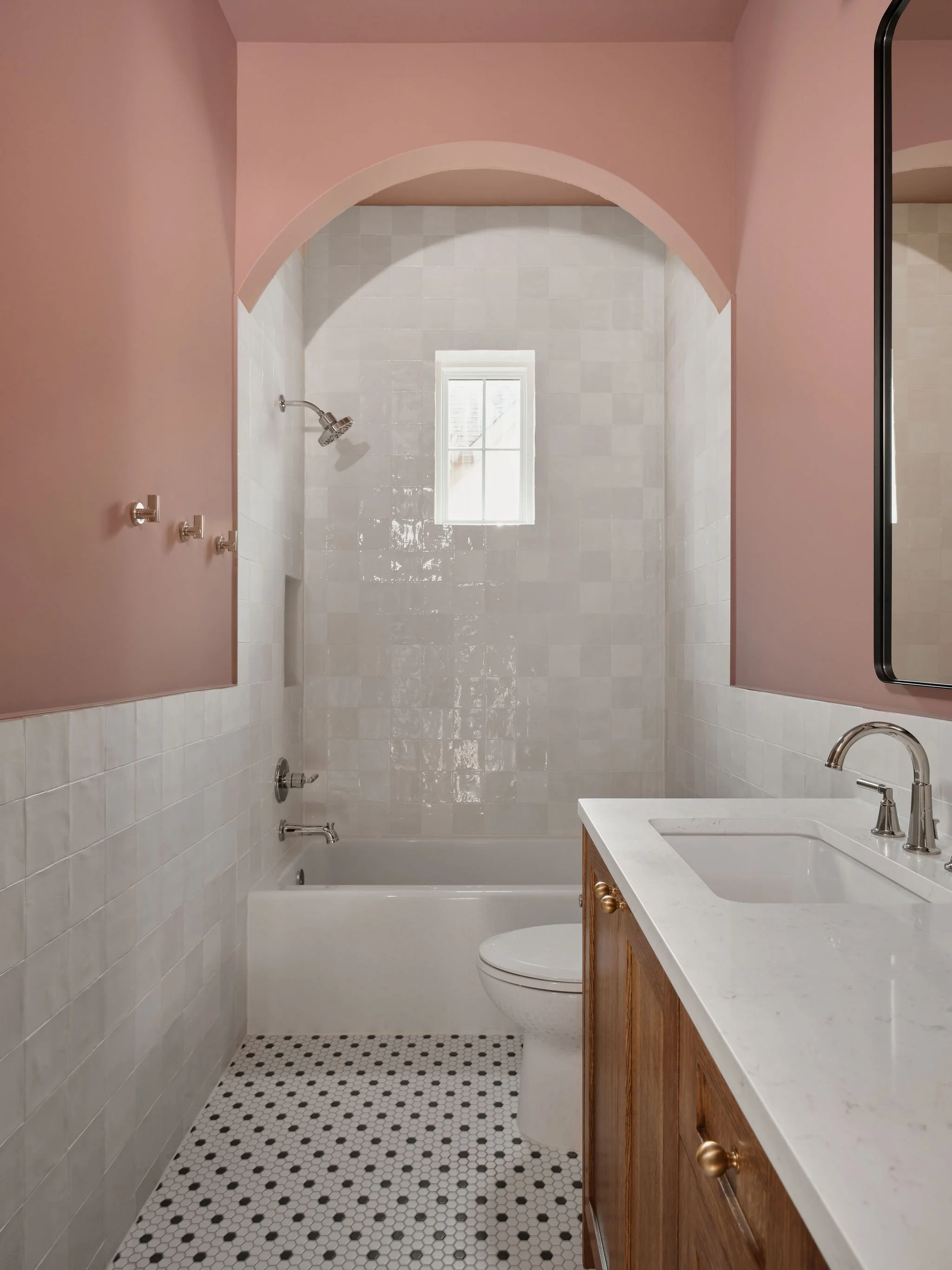 A small bathroom with pink upper walls, a white tile shower area with a window, a white toilet, and a wooden vanity with a white marble countertop and a sink.