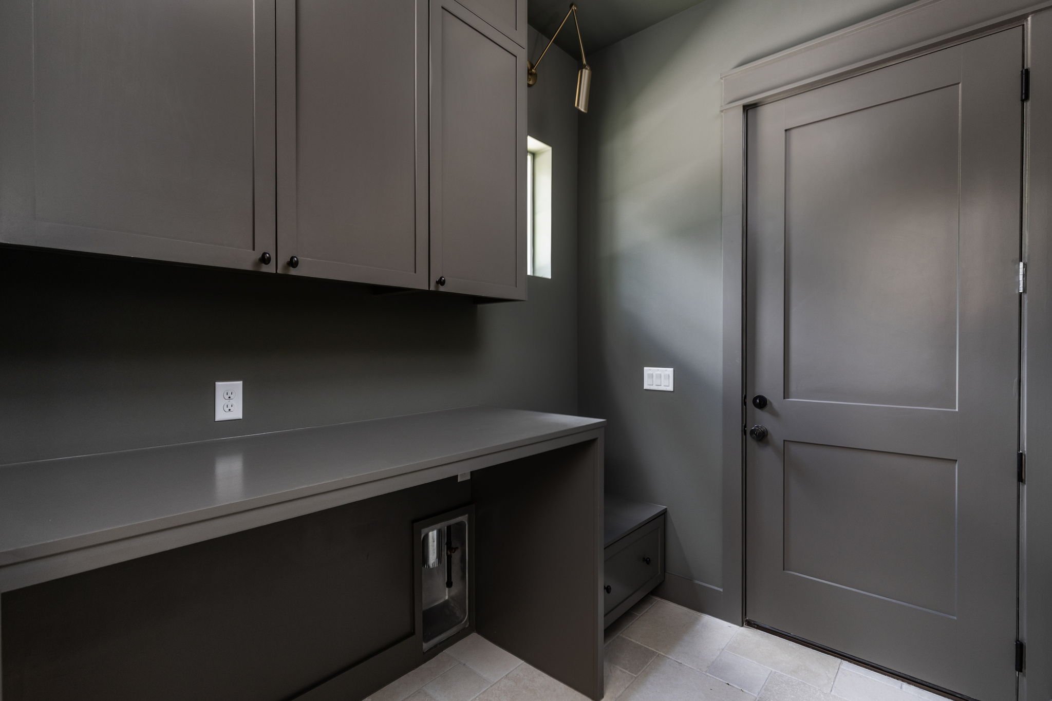 A small modern laundry room with gray cabinets, a gray countertop, a small window, and a gray door.