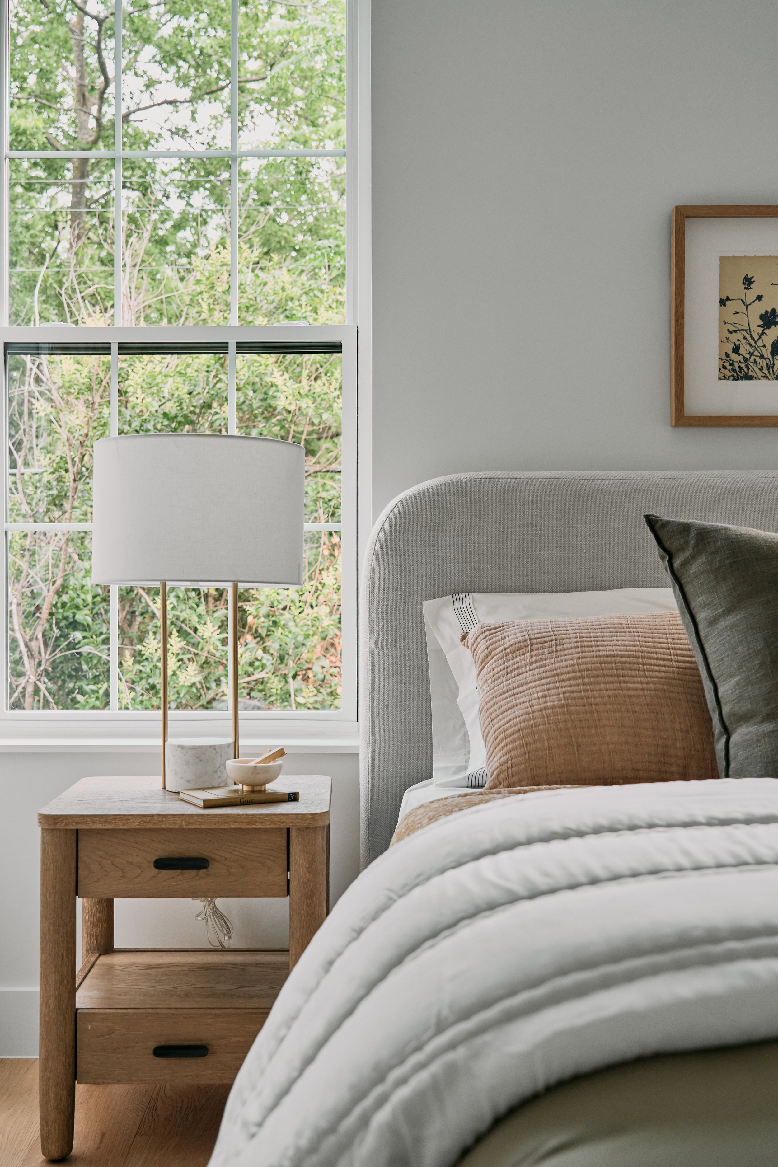 Minimalist bedroom with a bed, wooden nightstand, and lamp. The bed has a light gray headboard and is adorned with earth-toned pillows and a white comforter. A large window with a view of greenery is in the background. A framed artwork is hung on the