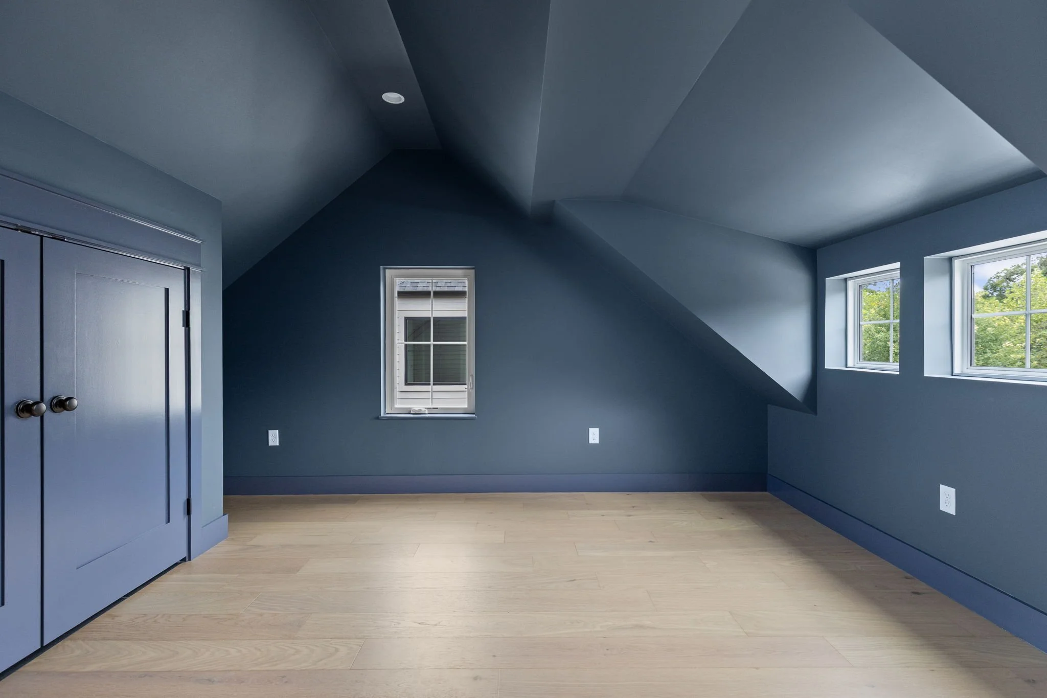 Empty blue room with two windows showing green trees outside, light wooden floor, closet with double doors, and a sloped ceiling with recessed lighting.