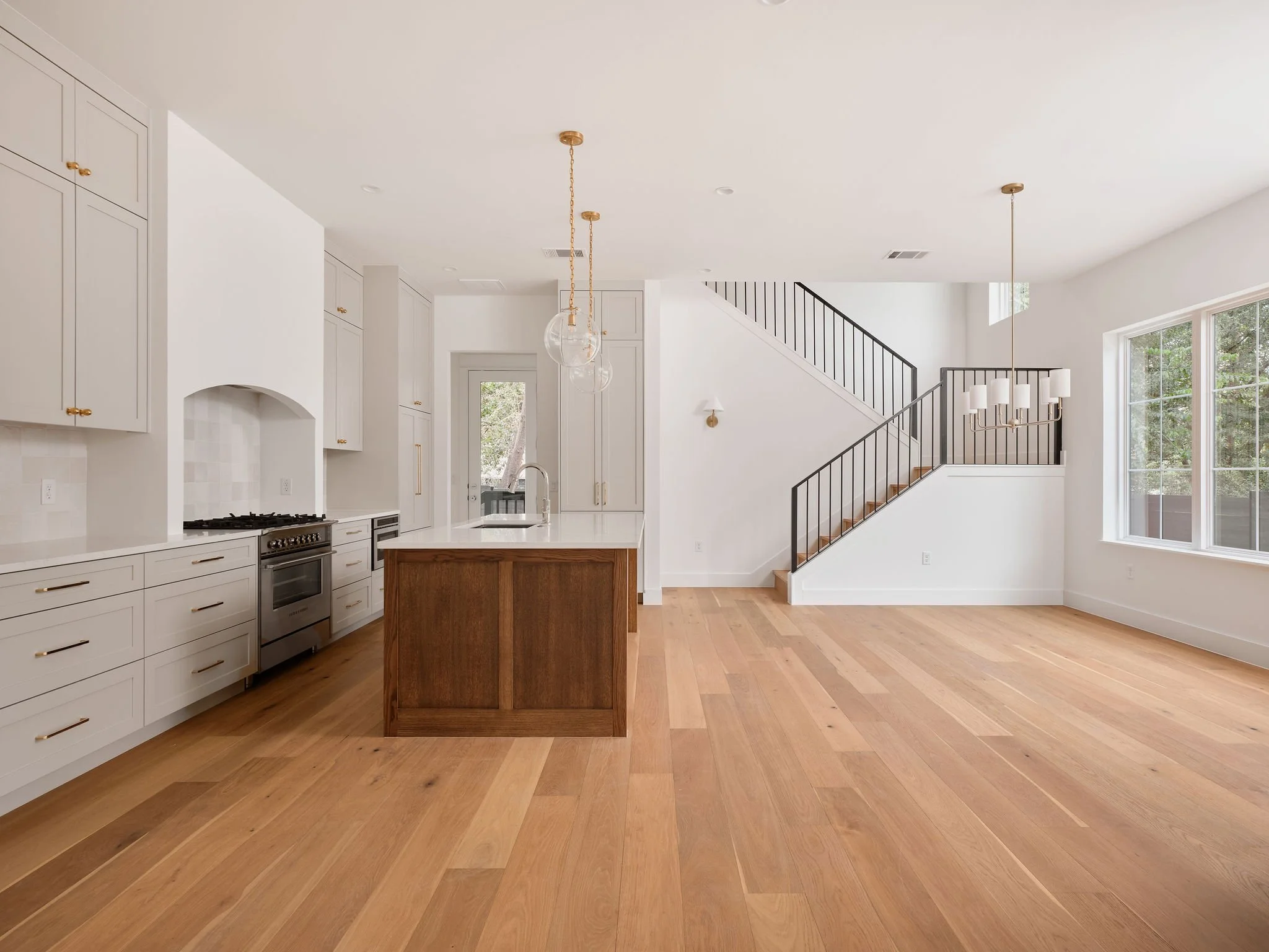 Empty modern kitchen and living area with hardwood floors, white cabinets, stainless steel stove, a kitchen island with white countertop and wooden base, large windows, and a staircase with black railing leading upstairs.