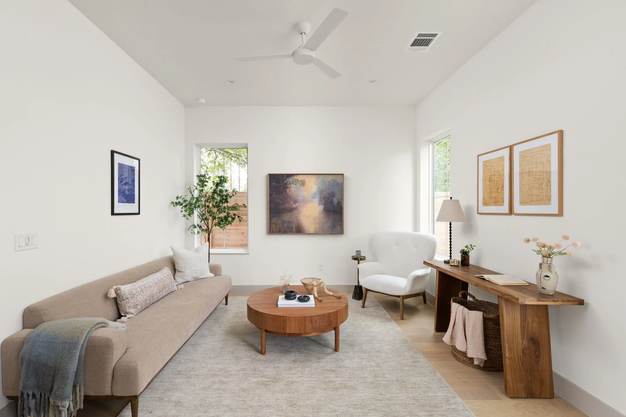 A bright, minimalist living room with white walls, a beige sofa, white armchair, wooden coffee table, and various wall arts, including a landscape painting and framed drawings. There's a window with greenery outside, a floor lamp, and a wooden side t