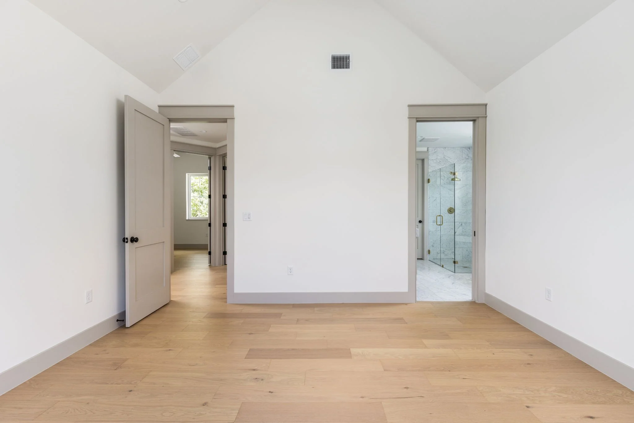 Empty room with wooden flooring, white walls, three doorways leading to other rooms, and a window with natural light.