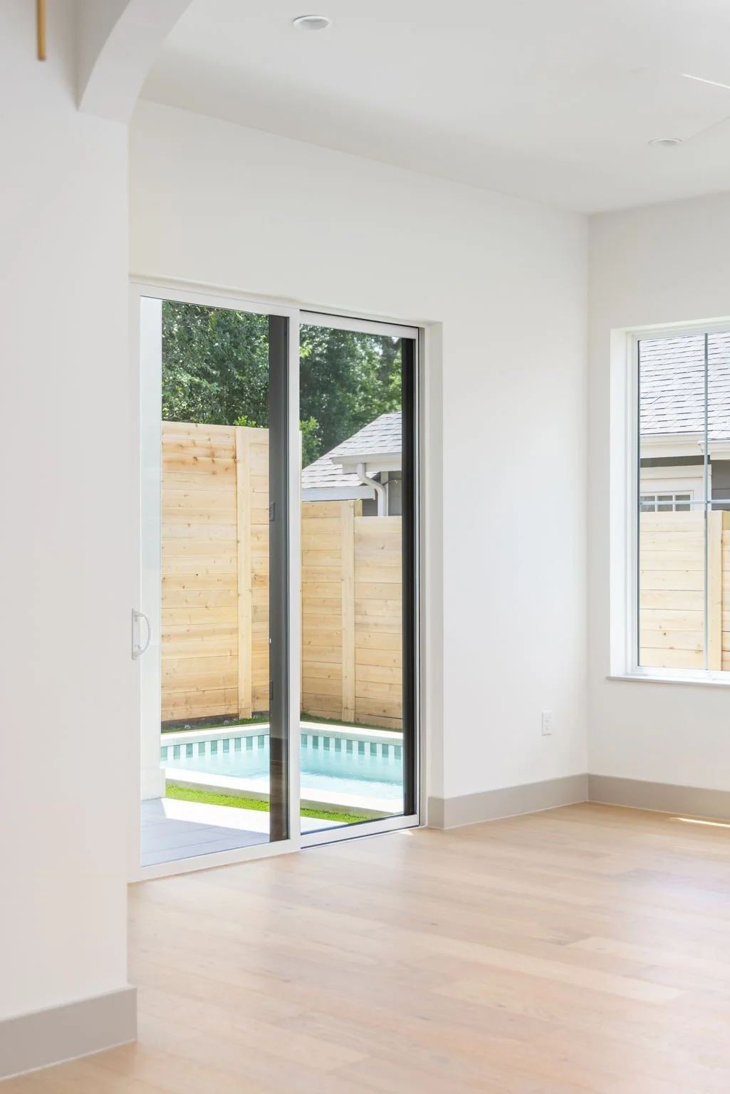 Empty room with light wood flooring, white walls, a sliding glass door leading outside to a small yard with a pool, and a window showing a neighboring house.