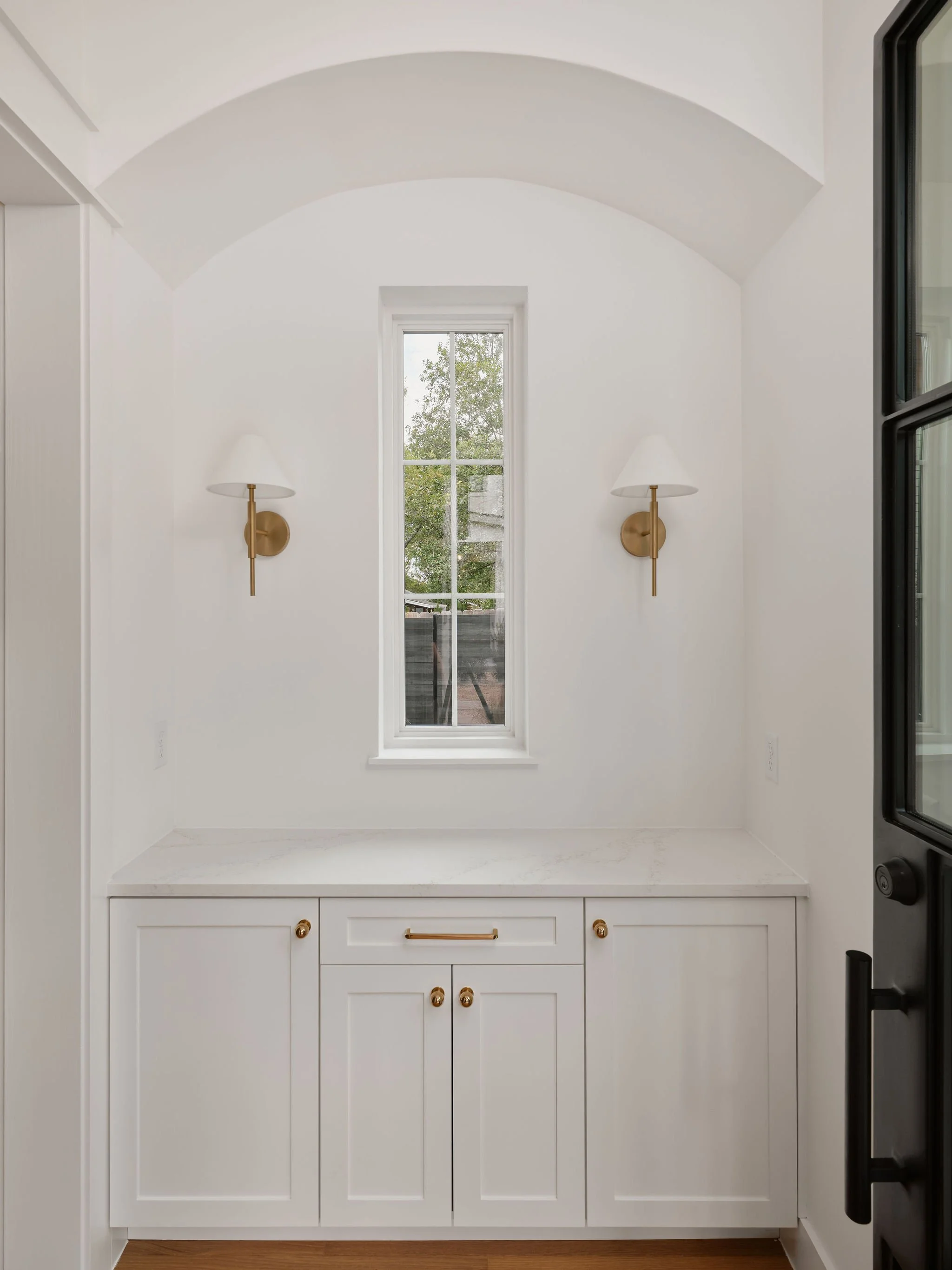 Bright white kitchen corner with a small window, two wall sconces, and a white cabinet with gold handles.