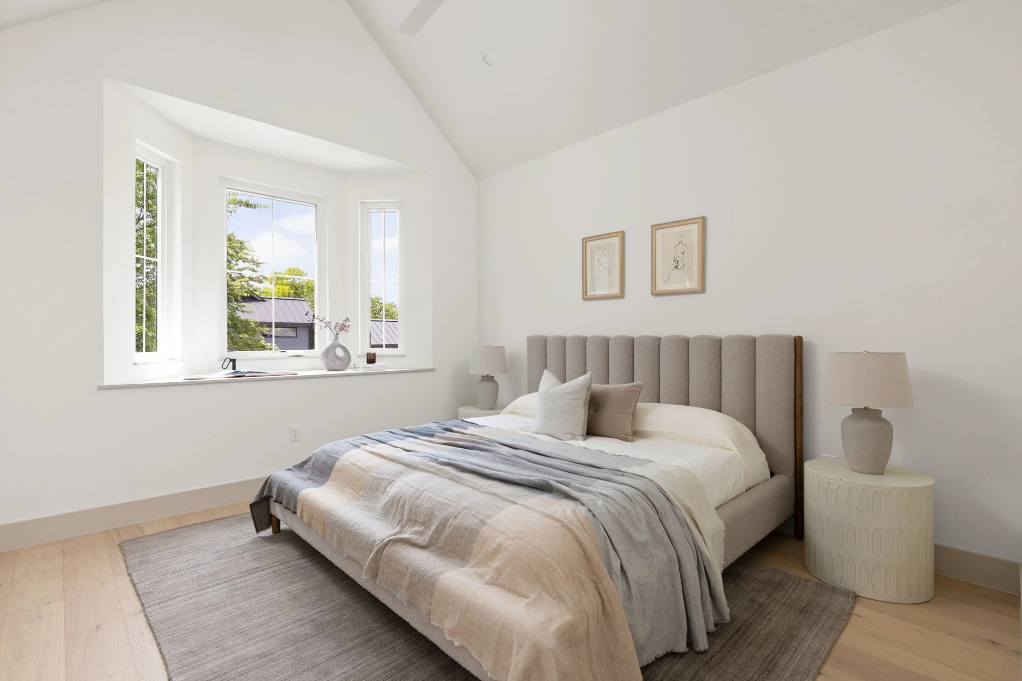A bedroom with a large bed, beige headboard, white bedding, and pillows. Two side tables with lamps, two framed artworks on the wall, a large window with plants outside, and a small vase with flowers on the windowsill.