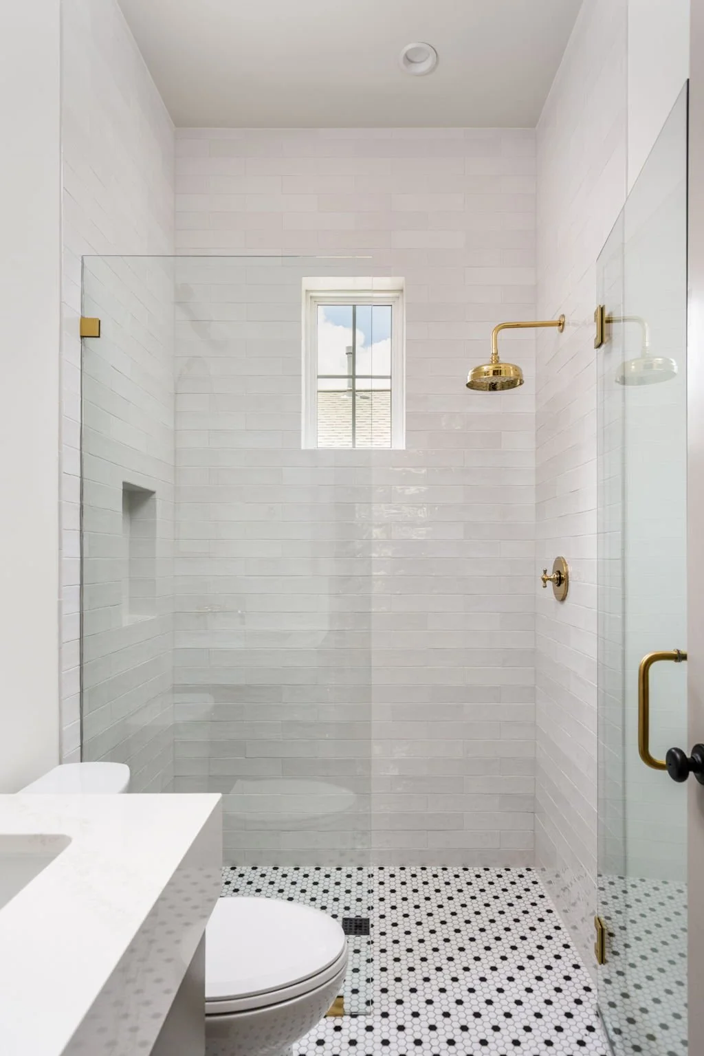 Bathroom with a walk-in shower enclosed by a glass door, brass fixtures, white subway tile walls, a small window, a white toilet, and a white sink with a marble countertop. The floor has black and white patterned tiles.