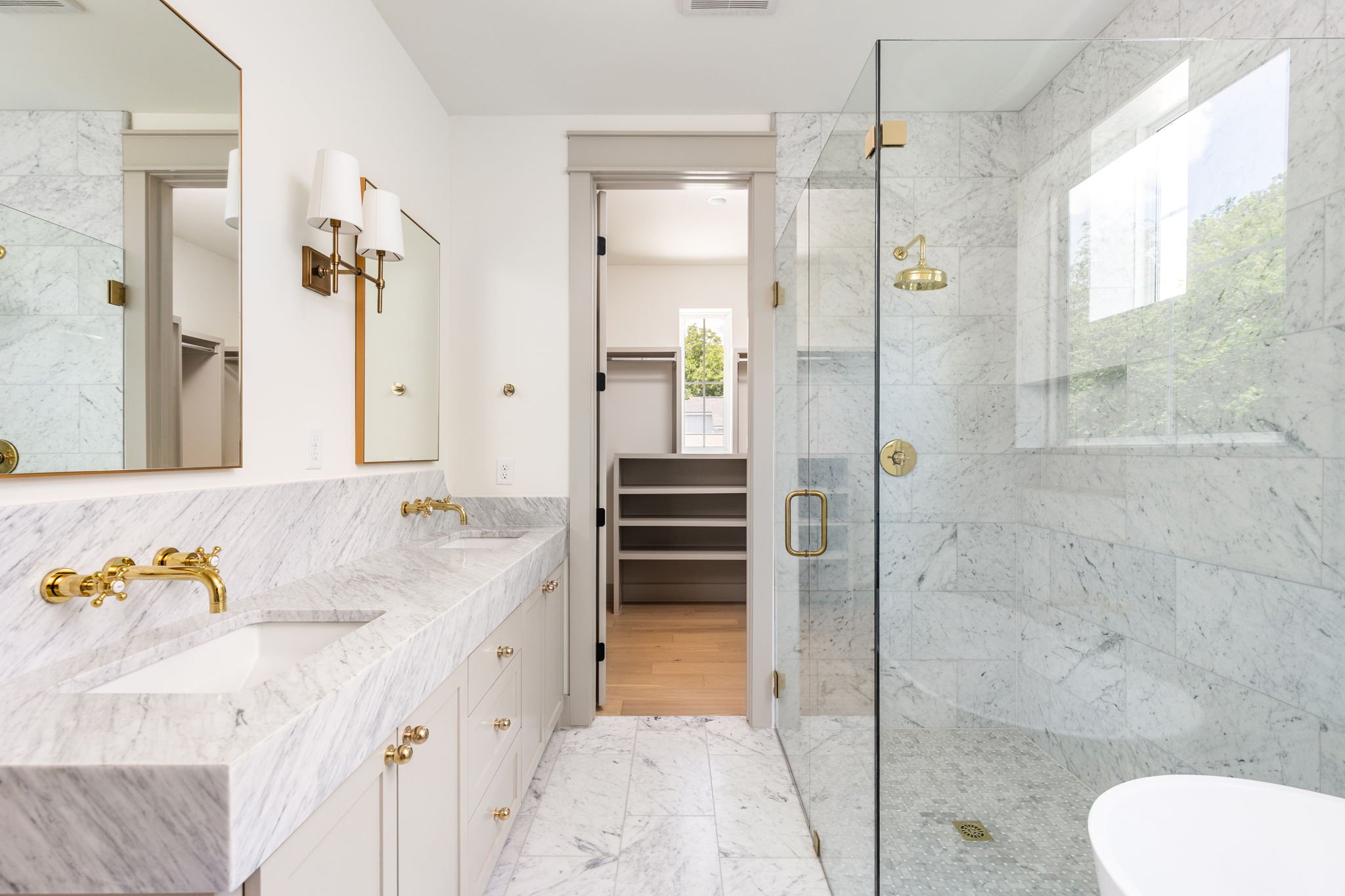 A marble bathroom with a vanity, gold fixtures, a large mirror, a walk-in shower with glass door, a window, and a small closet or storage space visible through an open door.