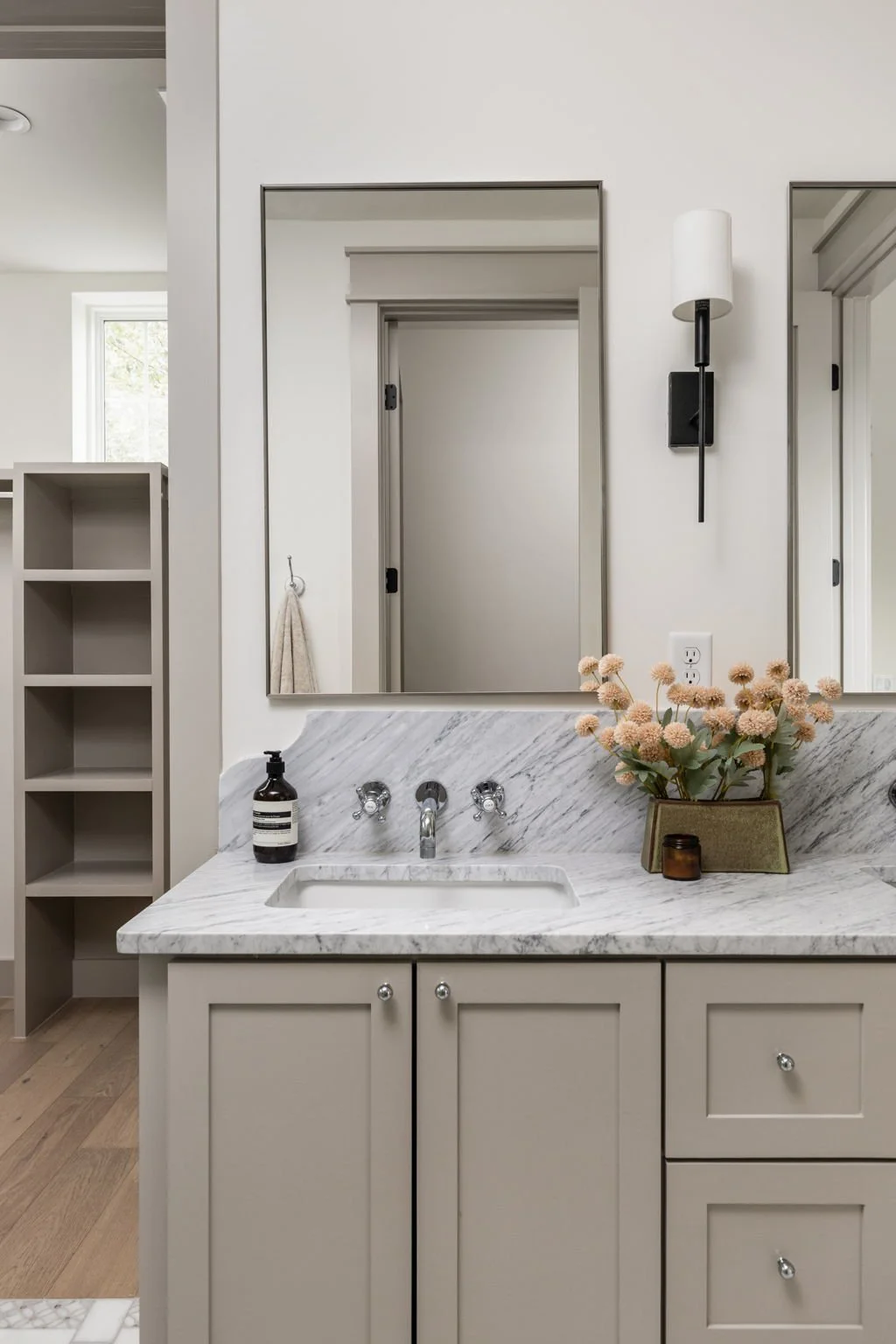 Modern bathroom with marble countertop, beige cabinetry, a mirror, wall-mounted light, flowers in a vase, and a soap dispenser.