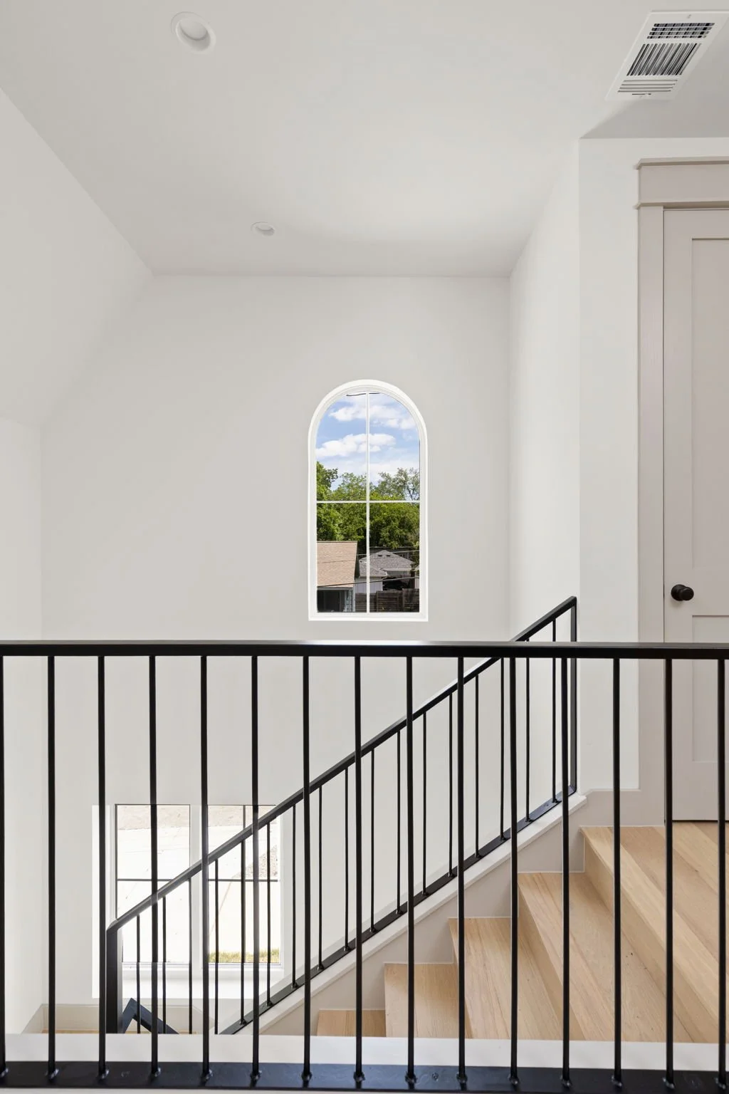 Interior view of a modern staircase in a house, featuring wooden steps, black metal railing, a white wall with an arched window showing trees and a cloudy sky, and a door on the right side.