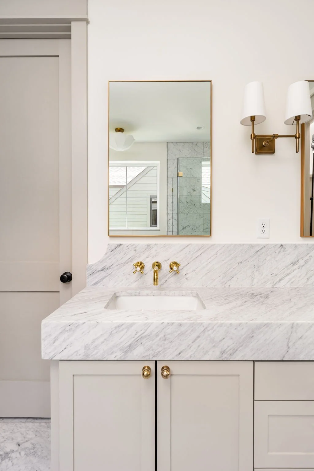 Bathroom vanity with white marble countertop, gold fixtures, and a mirror above, reflected in the mirror is a window and a shower with marble tile.