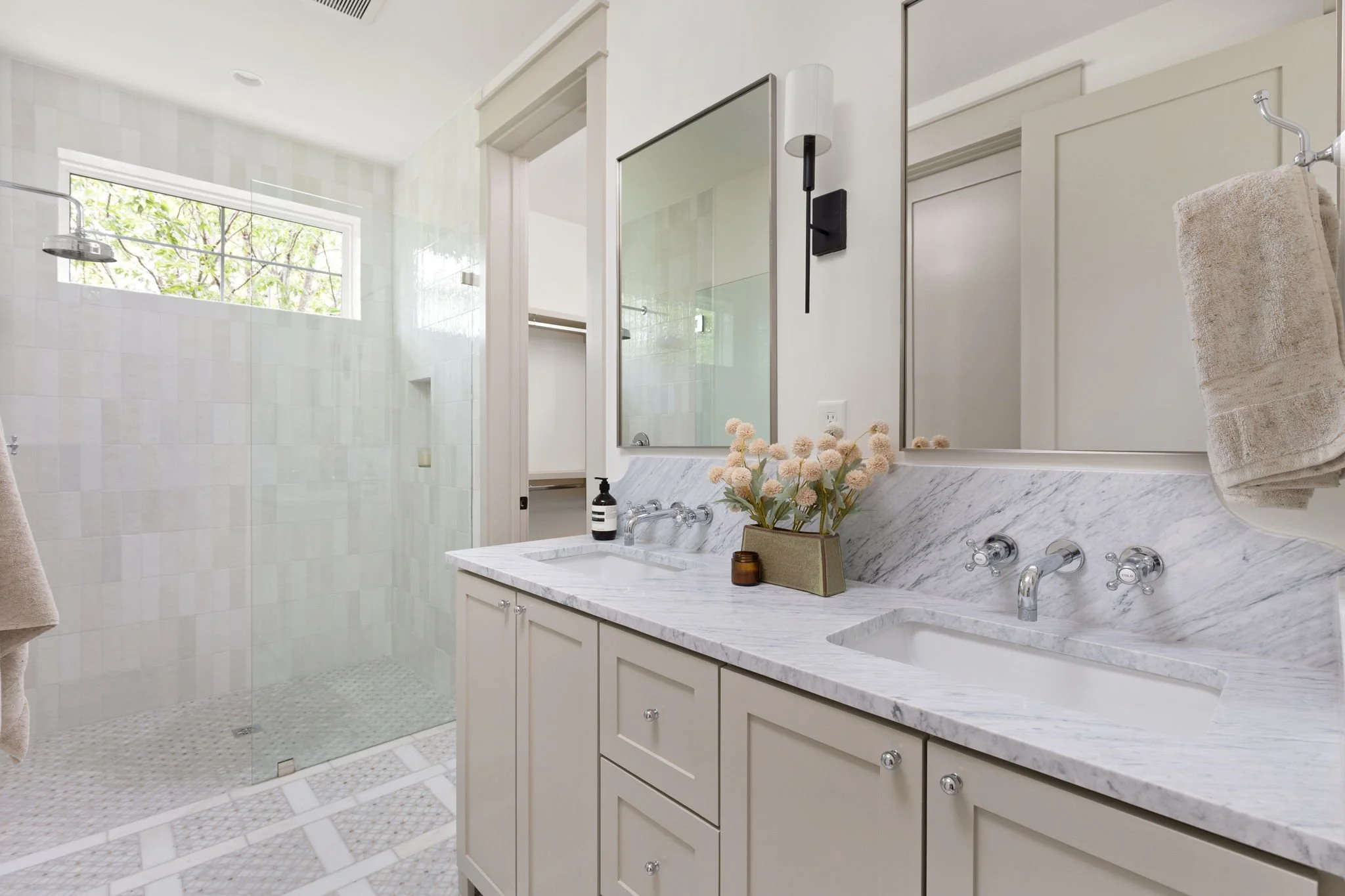 Modern bathroom with white cabinetry, marble countertop, and large mirror above sink; beige towel on hook, window with tree view, and walk-in shower with glass door and white tiled walls.