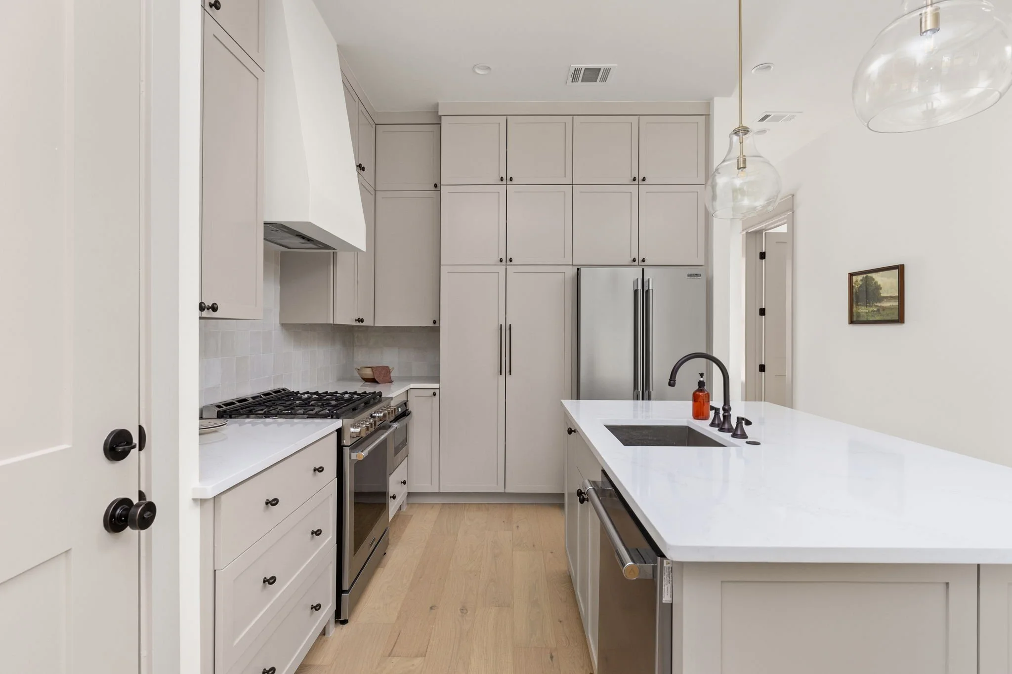 Modern white kitchen with cabinets, a stainless steel refrigerator, a gas oven, pendant lights, and a white kitchen island with a black sink and faucet.