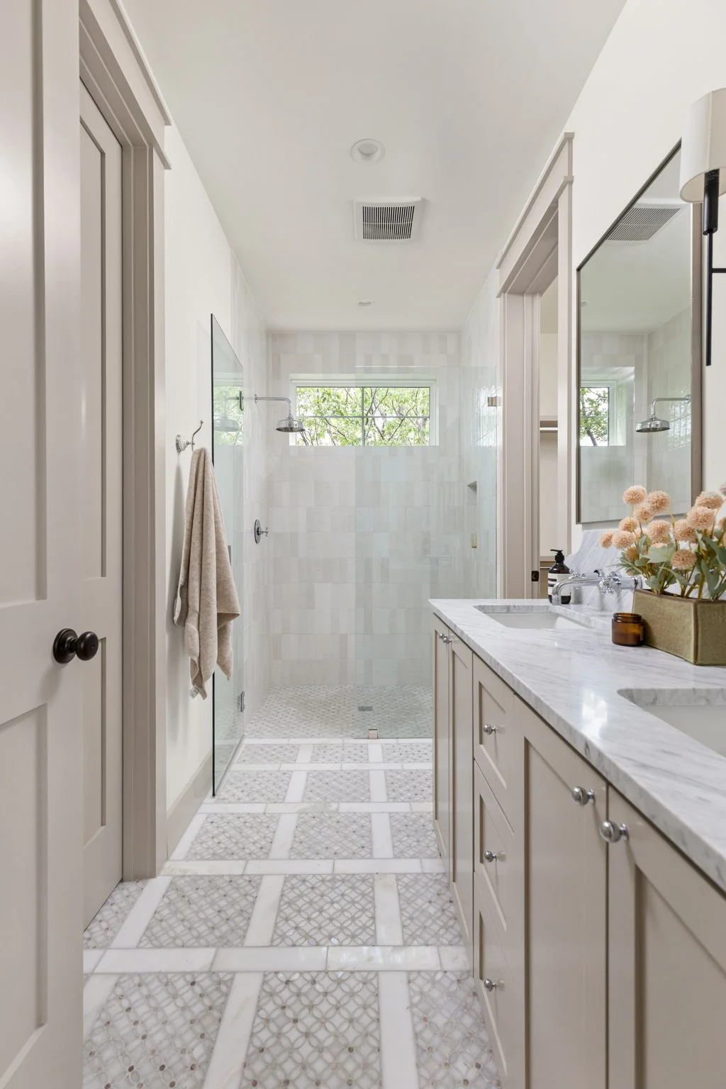 Bright bathroom with double sinks, marble countertop, large mirror, flower arrangement, walk-in shower with glass door, and small window with greenery outside.