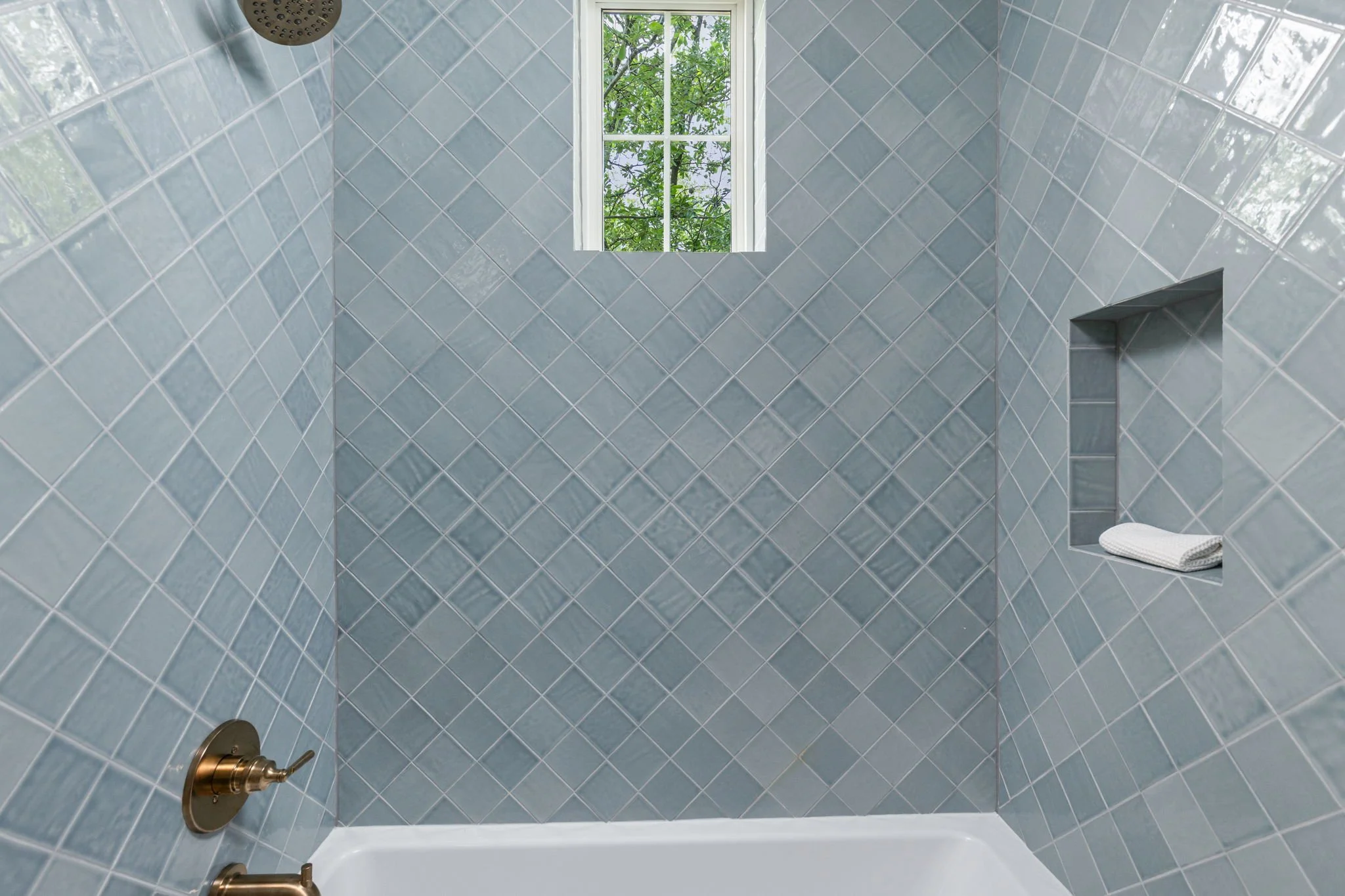 The image shows a shower with blue diamond-shaped tiles on the walls, a small window with a view of greenery outside, a showerhead at the top left corner, and a rolled white towel on a ledge.