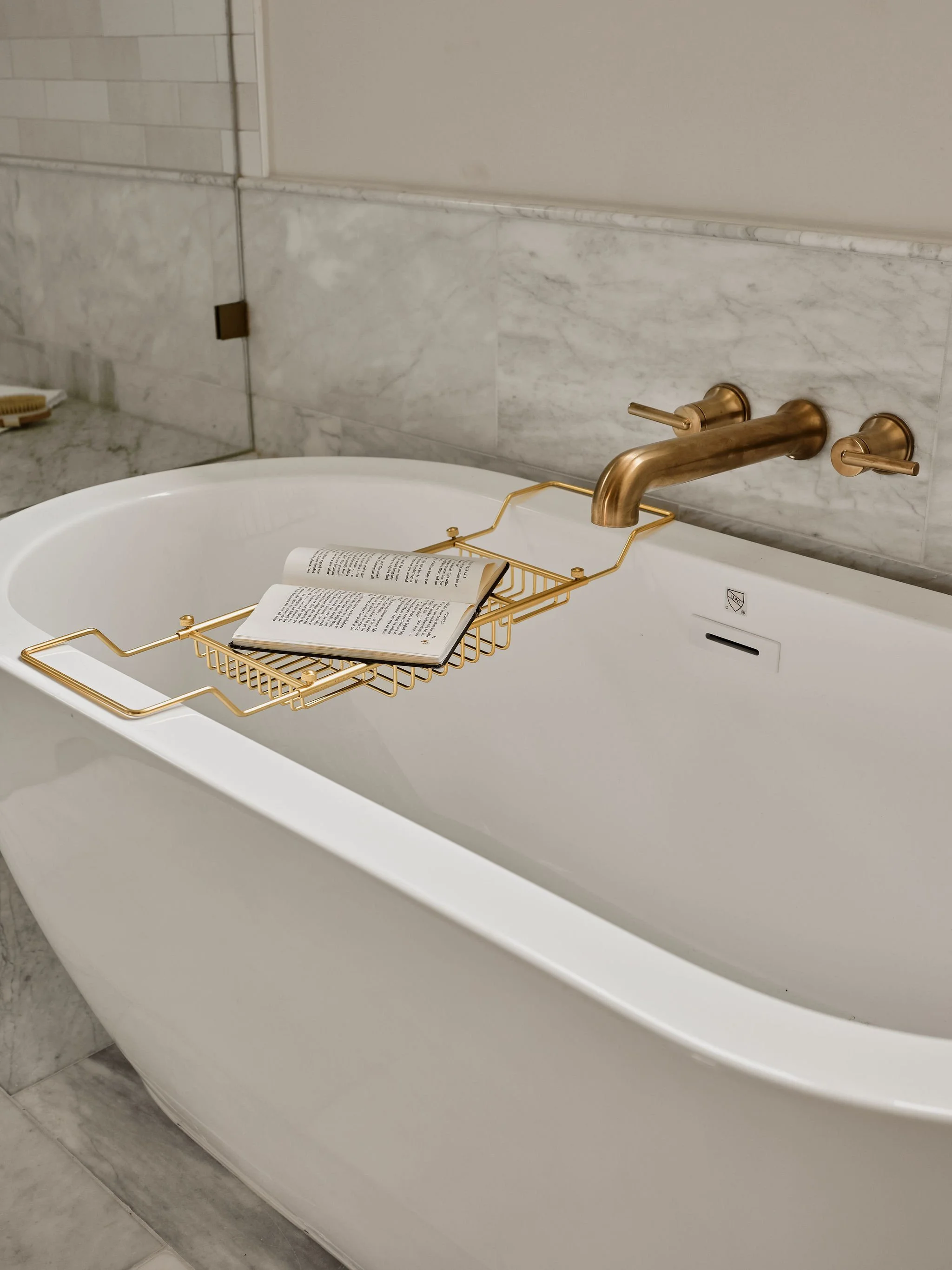 A white bathtub with a gold-colored metal bath tray holding an open book, set against a marble wall with a gold faucet and matching controls.